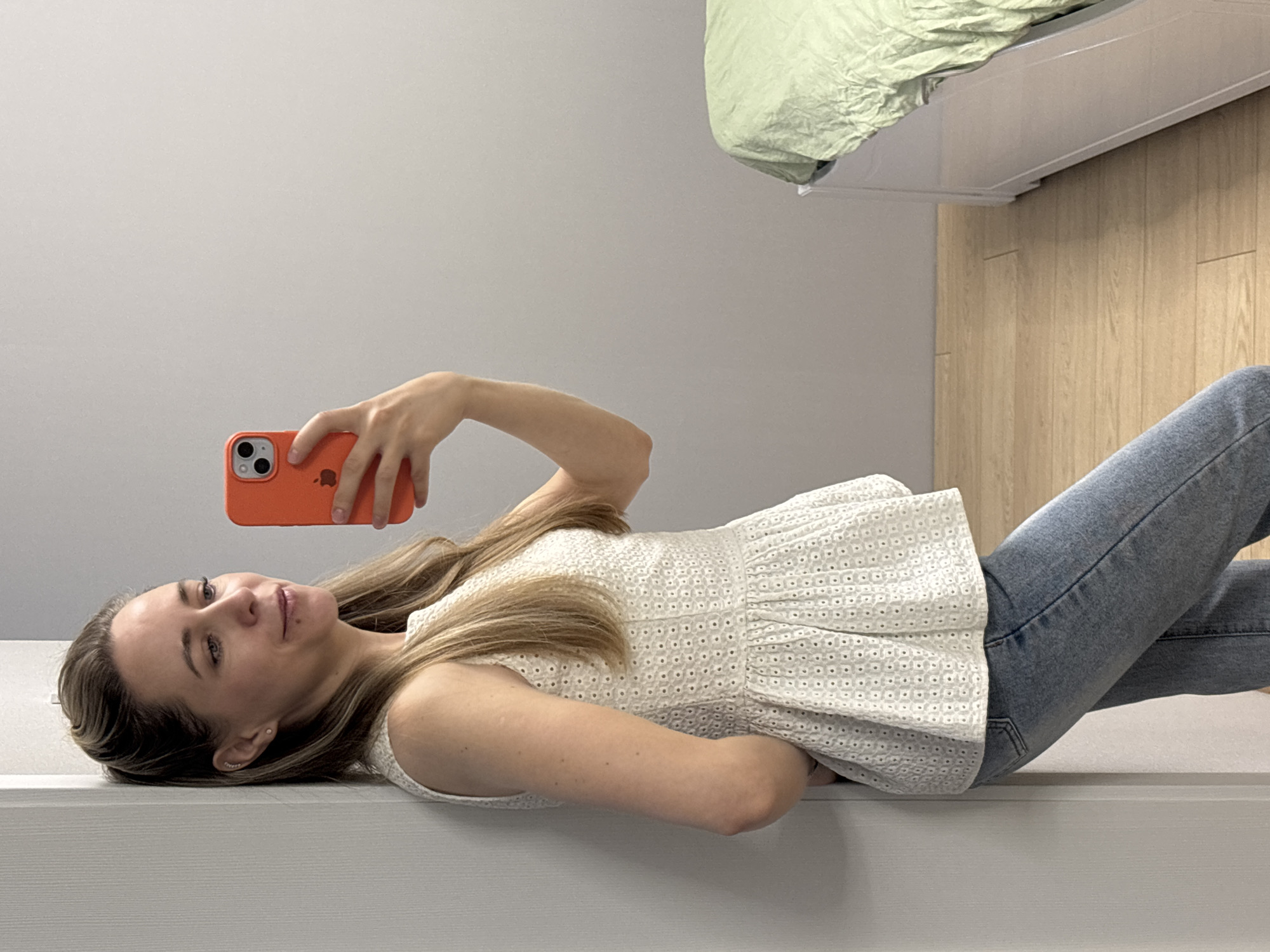 A young woman with long, light brown hair is depicted lying on her side indoors, taking a selfie with an orange smartphone. She is wearing a white sleeveless top with an eyelet pattern and light blue jeans. Her right hand holds the phone, which features a dual-camera system and an Apple logo on its back. Her expression is calm, with a slight smile as she looks towards the phone's camera. She is lying on a low, white platform or bench. In the background, a light grey wall meets a light-colored wooden floor, and a bed with a light green duvet is partially visible. The scene is brightly lit, typical of an indoor setting during the day. The location context provided is Chaikovsky, Russia.