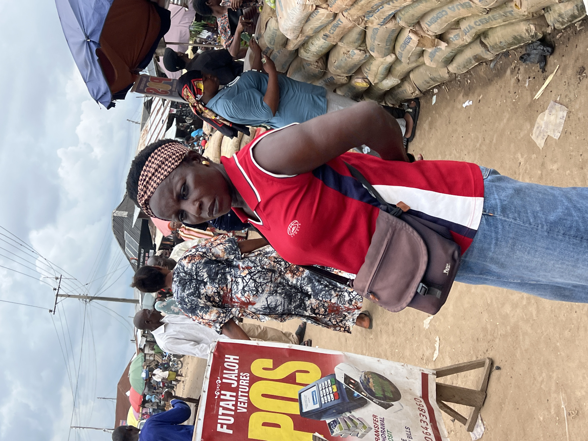 In the city of Asaba, Nigeria, a bustling marketplace scene unfolds under an overcast sky. The foreground is dominated by a woman wearing a red and white collared shirt and a denim skirt, with a brown crossbody bag slung over her shoulder. Her expression is serious as she looks to her left. To her left, a red and white sign advertises "FUTAH JALOH VENTURES" and "POS" services, featuring an image of a POS machine and stacked banknotes. Behind her, other individuals are visible, including a woman in a patterned dress, and further in the background, market stalls with various goods and umbrellas. To the right of the woman in red, sacks of cement are stacked high, forming a wall-like structure. The ground is dusty, with scattered debris suggesting an active commercial area. The overall impression is one of a lively, possibly chaotic, local market environment.