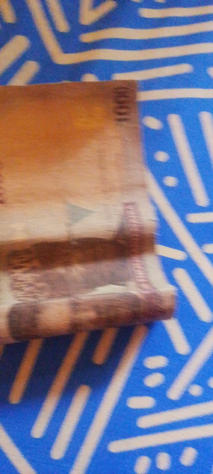 A close-up, slightly blurry, low-angle shot shows a stack of folded banknotes, appearing to be Nigerian Naira, resting on a blue fabric with a white abstract pattern. The banknotes are a light brown or tan color. On the top banknote, the number "1000" is visible vertically along the right edge. The fabric beneath the banknotes is a vibrant blue with thick white lines and scattered white oval shapes forming a repeating design. The lighting is diffused, and the focus is soft, giving the image an almost painterly quality. There is no indication of the specific location within Jalingo, Nigeria, or any people present in the scene.