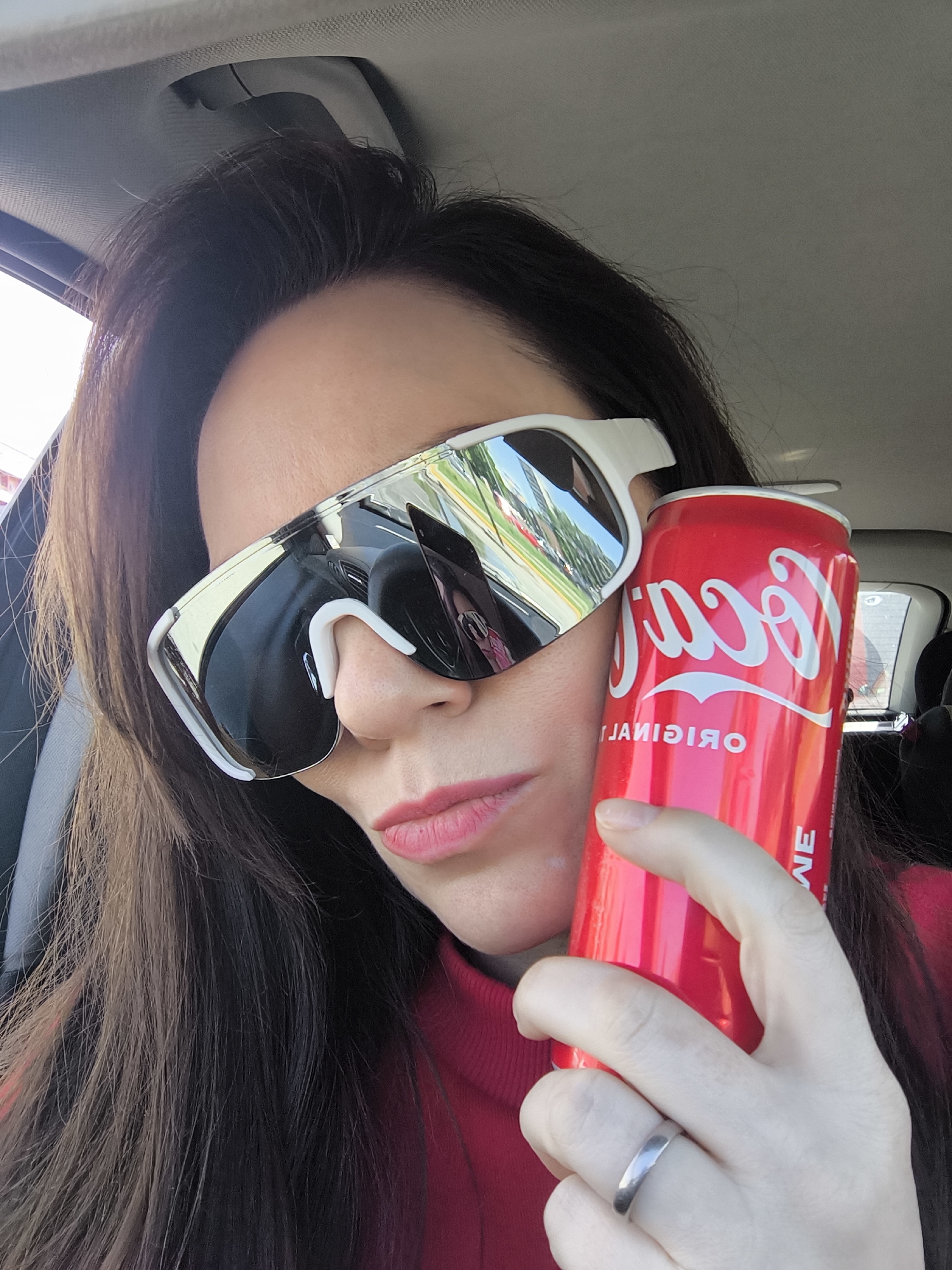 A woman with long, dark hair is in the front seat of a car, holding a red Coca-Cola can. She is wearing white-framed sunglasses that reflect buildings and trees, suggesting an urban setting. The reflection hints at architectural elements typical of Riga, Latvia, such as Art Nouveau buildings. The woman is wearing a red top. She has a ring on her finger. The car's interior is visible.
