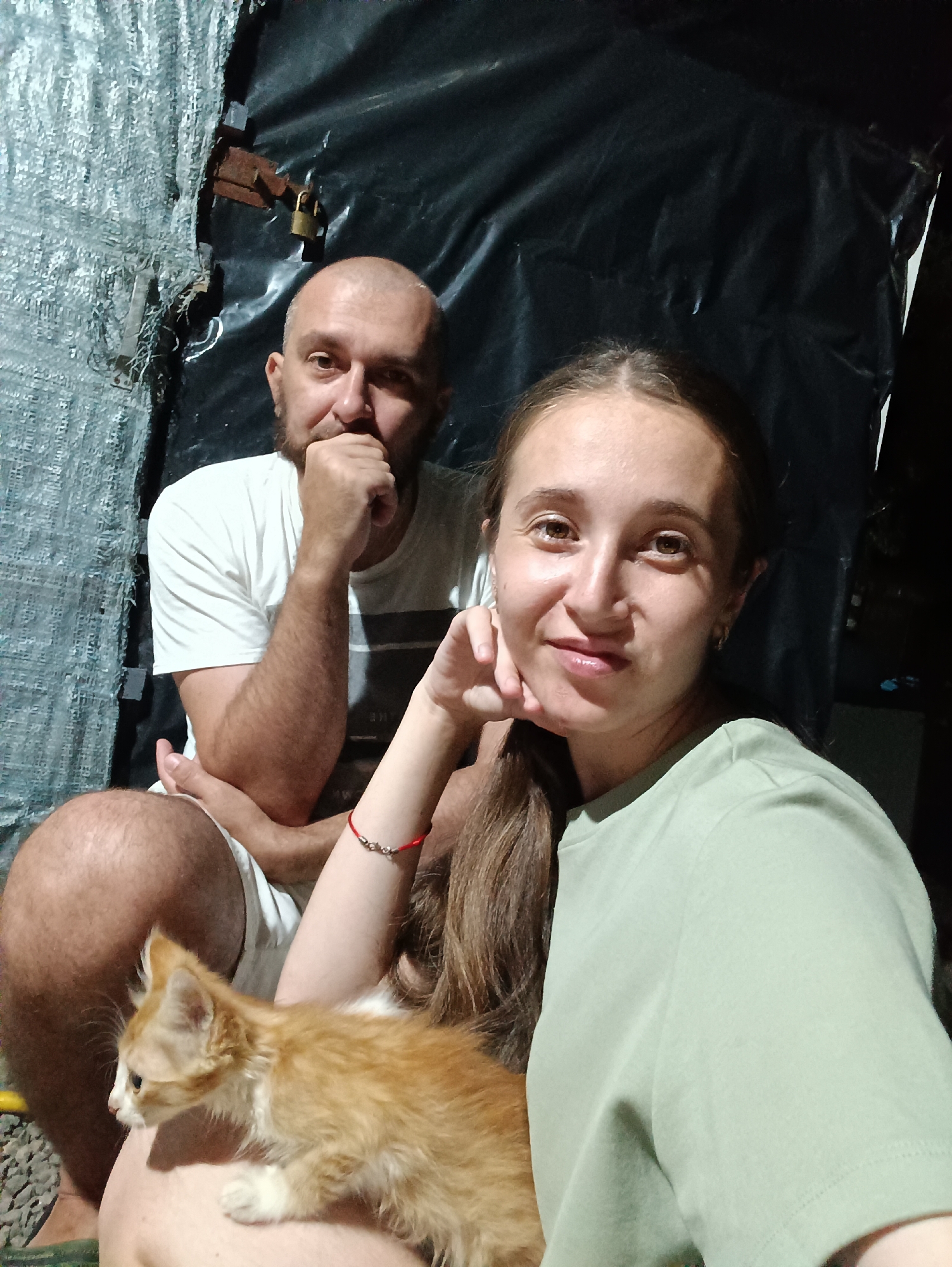 In the city of Vypasne, Ukraine, a man and a woman are seated outdoors with a ginger kitten. The man, bald with a beard, wears a white t-shirt and white shorts. The woman, with long brown hair, is dressed in a light green t-shirt. They are positioned close together, with the kitten resting on the woman's lap. The background consists of a black plastic sheeting, with a grey, textured material on the left. A rusty padlock is visible on the sheeting, attached to a metal fixture. The lighting is dim, suggesting it is either dusk or nighttime.