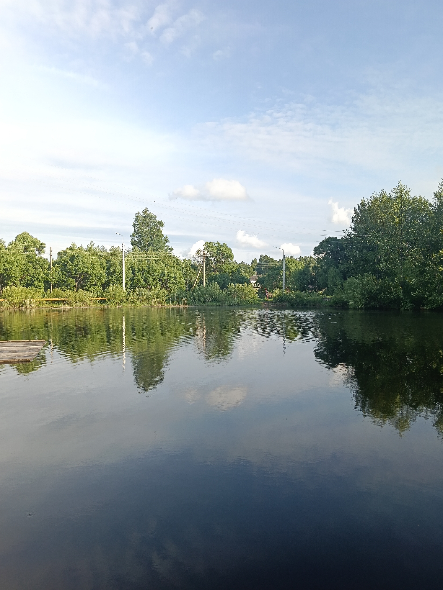 The image depicts a body of water reflecting the sky and surrounding vegetation. In the background, there's a line of trees and bushes, with utility poles visible amongst them. A small wooden structure extends into the water on the left side of the frame. The sky is light blue with scattered clouds. The water surface is mostly dark, with ripples and reflections of the sky and trees. The image was taken in the city of Красный Маяк, Russia.