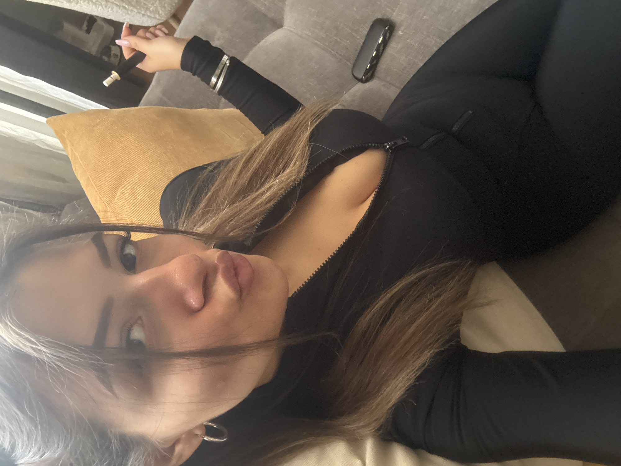 A woman is lying on a grey tufted sofa. She is wearing a black, long-sleeved, form-fitting jumpsuit with a zipper down the front. She is holding a black and silver vape pen in her right hand. A black electronic device is on the sofa near her. The background includes a window with blinds and a golden-colored pillow. The location is unknown and cannot be determined to be Sofia, Bulgaria.