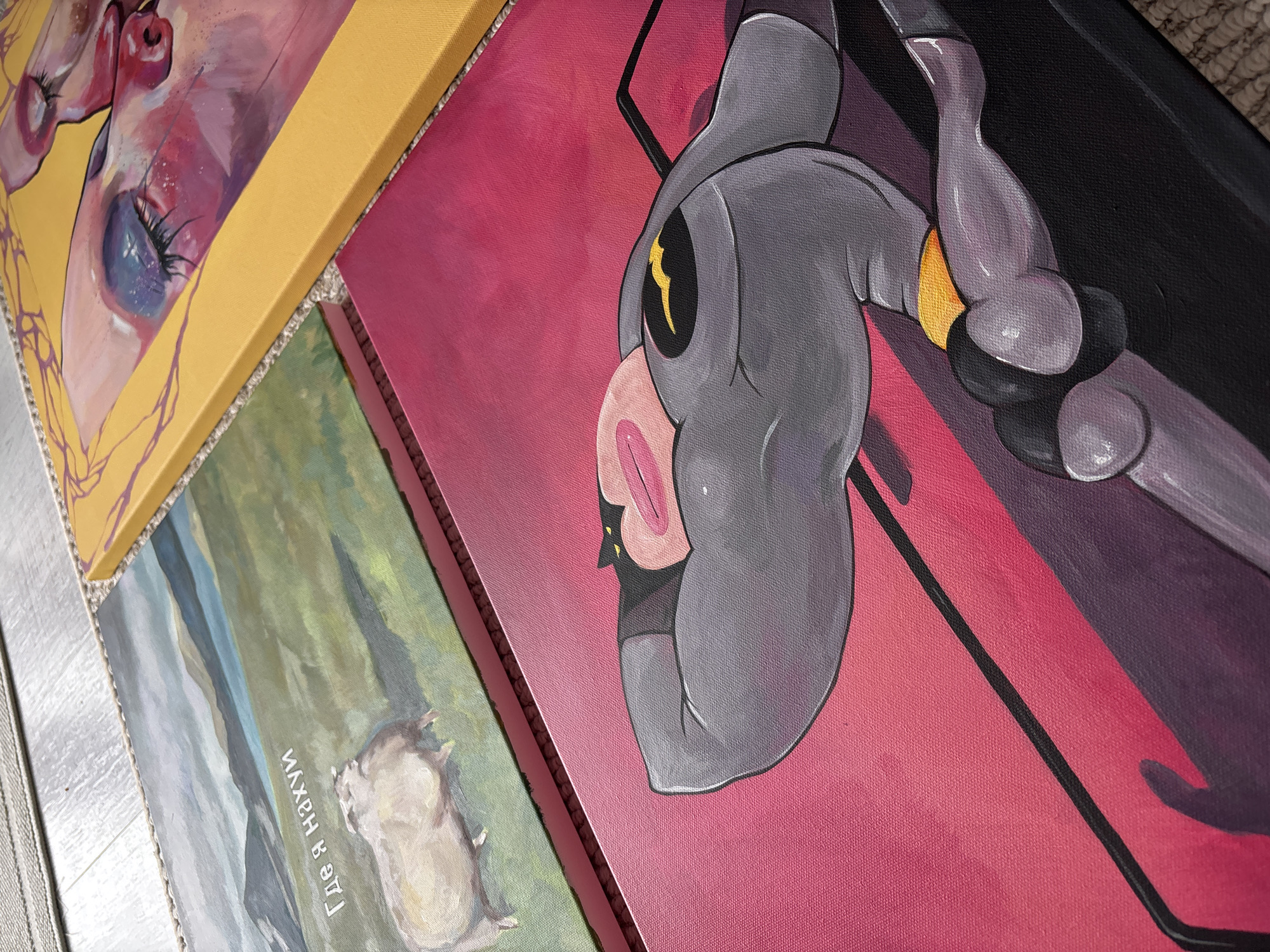 The image shows a collection of paintings on canvases. One painting is a portrait of a person with exaggerated features, predominantly in yellow and purple tones. Another painting depicts a landscape with an animal in the foreground, likely a rodent, with text in Russian that appears to read "Где я Нахлы." A third canvas features a cartoon-style character in shades of grey with yellow accents, against a pink and black background. The paintings are arranged close together. There is no information in the image to suggest a location in Krasnoyarsk, Russia.