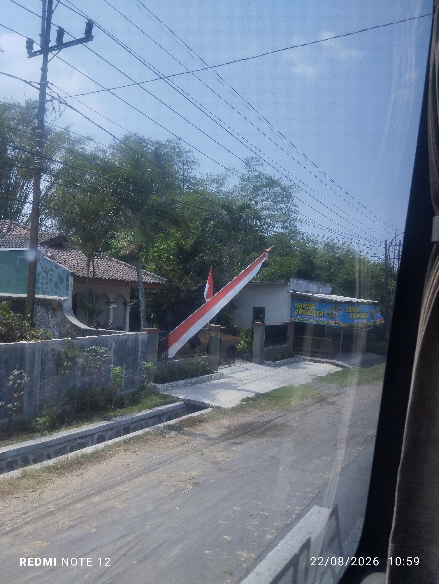 The image shows a street scene in Klagen Gambiran, Indonesia. A red and white Indonesian flag is prominently displayed, draped diagonally across the frame. To the left, a house with a tiled roof and a light-colored stone facade is visible, bordered by a concrete wall and some greenery. On the right, a small building with a blue banner advertising "BAKSO ANGKASA" (a type of meatball soup) is situated. Power lines and utility poles crisscross the sky, with lush trees and vegetation forming the background. The foreground consists of a dirt road and a concrete drainage channel. The timestamp "22/08/2026 10:59" and "REDMI NOTE 12" are watermarked at the bottom.