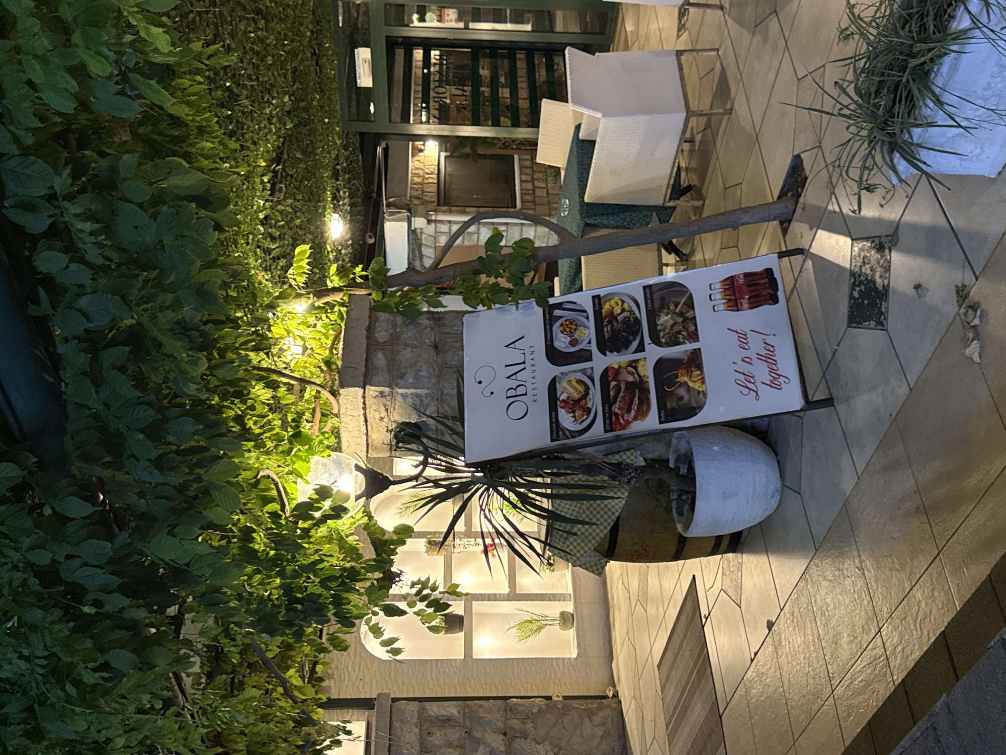 This image depicts an outdoor seating area of a restaurant called "OBALA RESTAURANT" in Budva, Montenegro. The scene is illuminated by ambient lighting, casting a warm glow. In the foreground, a white A-frame sign displays menu items with pictures, featuring dishes such as "Cevapi" and "Pasta," and a slogan "Let's eat together!". Two bottles of Coca-Cola are visible on the sign.
To the left of the sign, a large planter pot contains a spiky plant, and behind it, a wooden barrel is partially visible. Further back, a building features illuminated niches displaying potted plants. The seating area itself includes a green tablecloth-covered table surrounded by white wicker chairs. A glass ashtray rests on the table.
The floor is paved with irregularly shaped tiles. Overhead, dense green foliage creates a canopy, with string lights interwoven through the leaves. In the background, the entrance to the restaurant is visible, with a green door and windows, and a sign indicating "KLIMATIZOVANO" (air-conditioned in Croatian/Serbian). The overall atmosphere suggests a pleasant evening dining experience.
