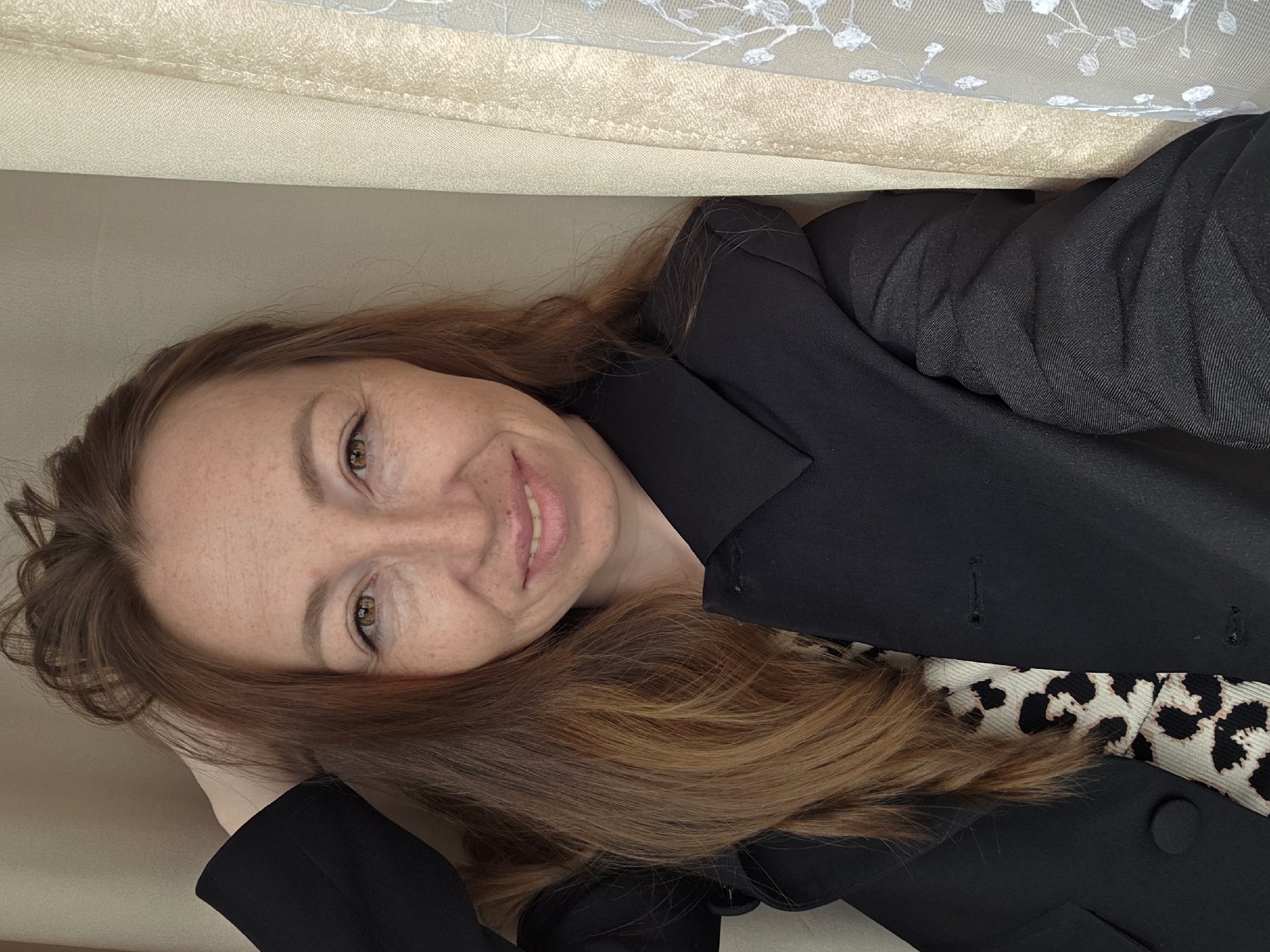 A woman with light skin, freckles, and brown hair is lying down, smiling at the camera. She is wearing a black blazer over a top with a leopard print pattern. She appears to be indoors, with a textured beige surface above her head. The location is specified as the city of Chaikovsky, Russia.