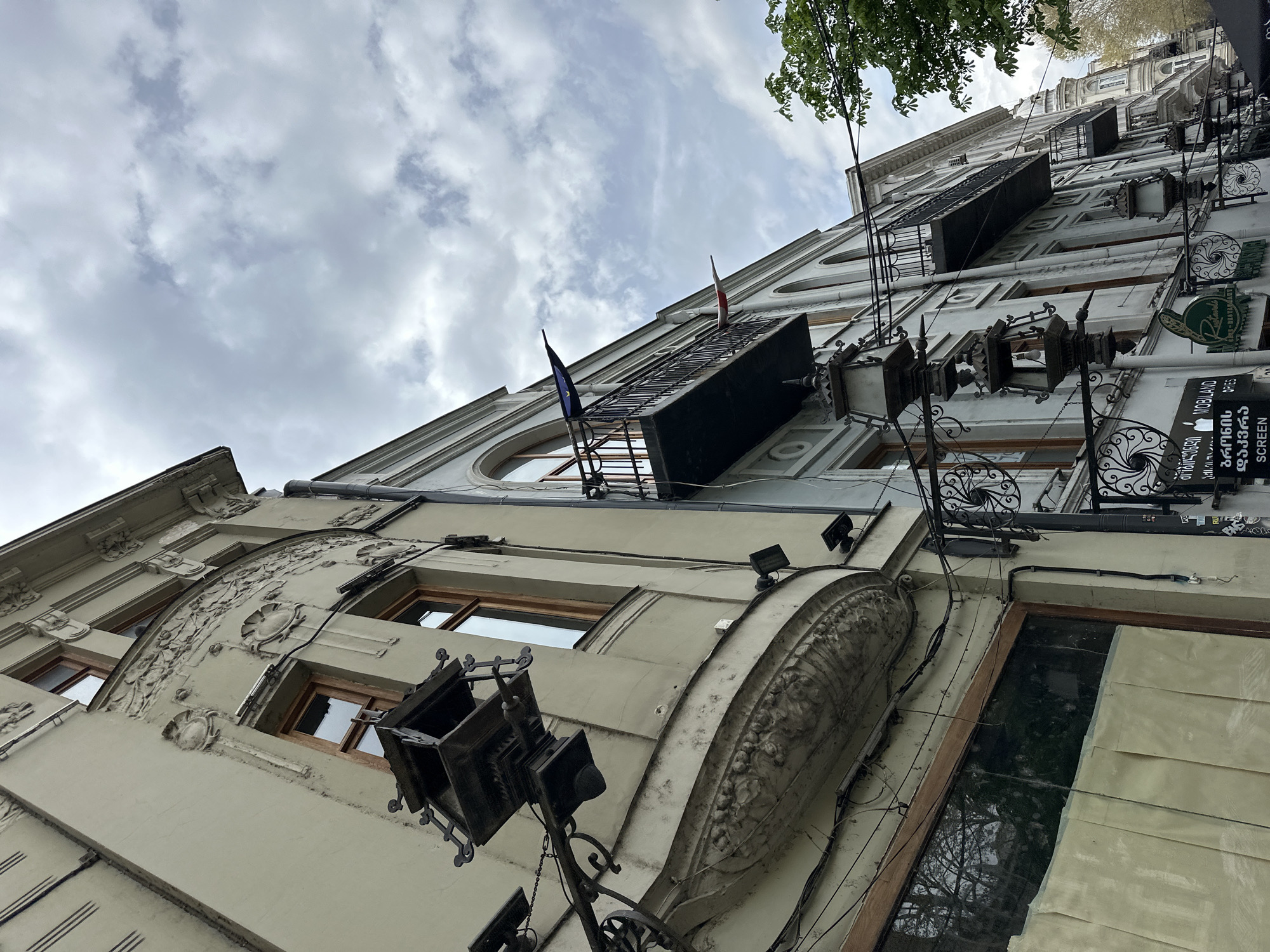 The photograph shows a low-angle view of an ornate building facade in Tbilisi, Georgia. The building is multi-storied with decorative moldings and architectural details, including curved window frames and sculpted elements. A small balcony extends from one of the upper floors, adorned with the flags of Georgia and the European Union. Ornate street lamps are attached to the building's exterior, and various signs are visible, including one in Georgian script and another with English text indicating a mobile device screen repair service. The sky is overcast with cloudy conditions. The composition emphasizes the verticality of the building and its historic architectural features.