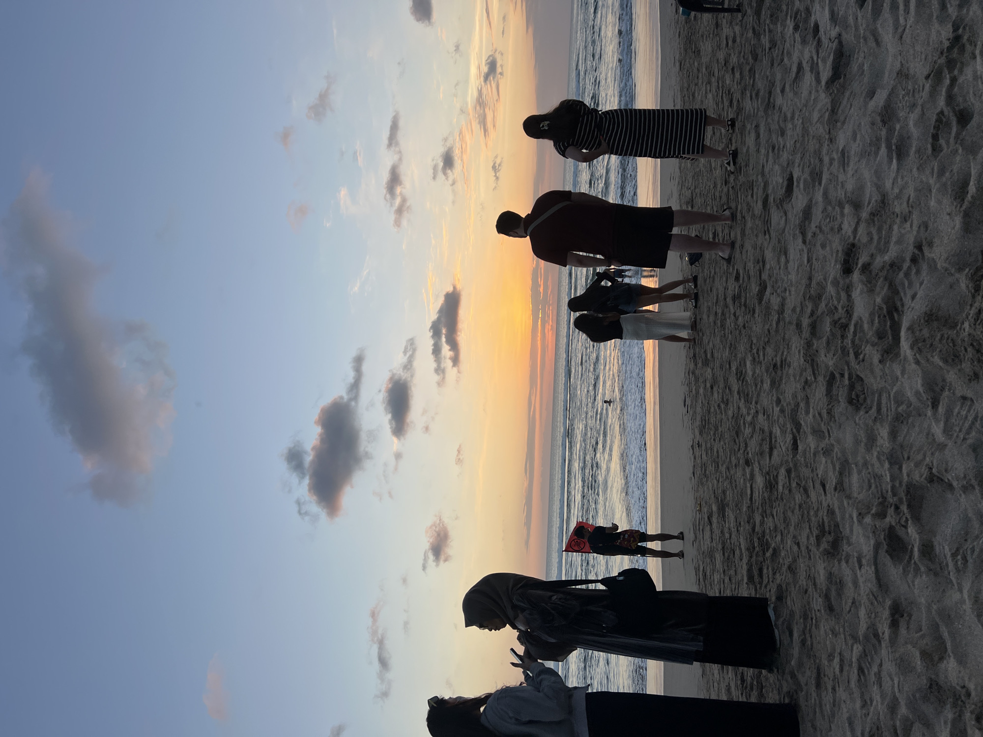 At sunset on Kuta Beach, Indonesia, several people are silhouetted against the sky. In the foreground, two women are talking. To their right, a man holding a red flag stands near the water's edge. Further to the right, a couple stands facing the ocean, and another woman in a striped dress is looking away from the camera. The sky is filled with pastel clouds, and the sun is setting over the calm ocean. The sand on the beach is textured with footprints.