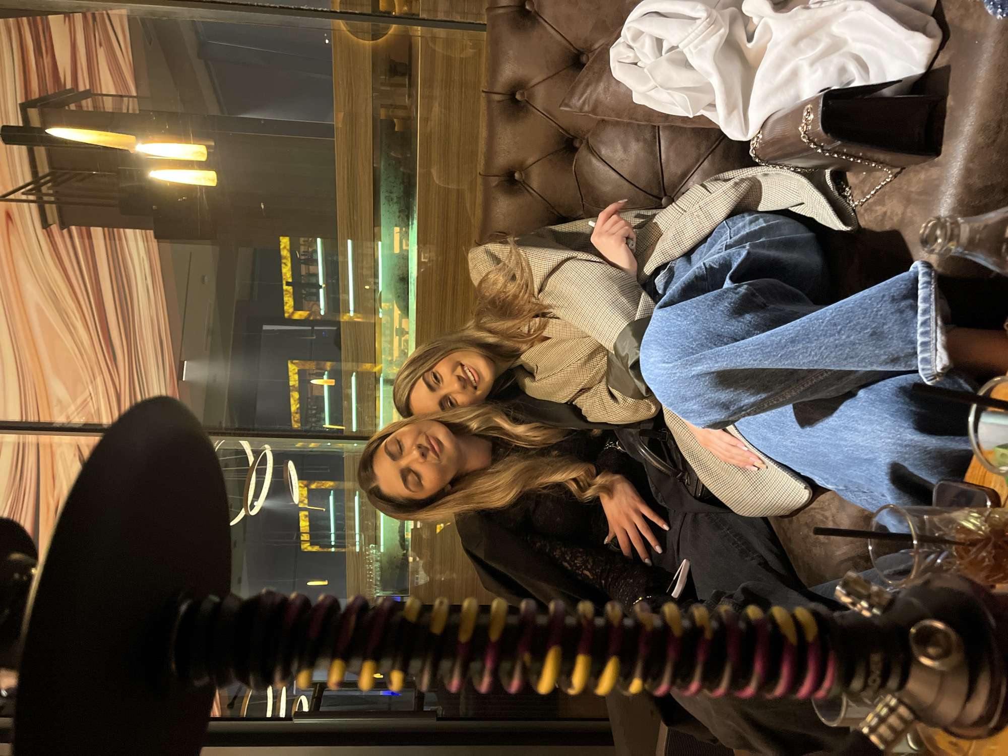The image depicts two young women relaxing and socializing in an upscale indoor lounge or bar, likely during the evening in Burgas, Bulgaria. They are seated on a rich brown, tufted leather couch.
The woman in the foreground on the left has long, wavy blonde hair and is dressed in a black top, looking towards the viewer with a serene expression. Her left hand is resting gently on her midsection. Behind her and to the right is another woman with long, light brown hair, wearing a stylish light-colored plaid blazer over a dark top and blue jeans. She is smiling cheerfully and gazing directly at the camera.
In the immediate foreground, a black hookah is partially visible, featuring a distinctive coiled hose with red, purple, and yellow sections. The word "ROVER" is discernibly printed on the black handle of the hookah hose connector. On the couch beside the women, a white folded garment and a dark brown or black handbag with a silver chain strap are placed. To the right, glimpses of glasses with straws and possibly empty bottles suggest the presence of beverages.
The background features large glass surfaces reflecting modern interior lighting, creating a sophisticated ambiance. The setting appears contemporary and well-appointed. Both women convey a sense of happiness and relaxation, enjoying their time together in this social environment.