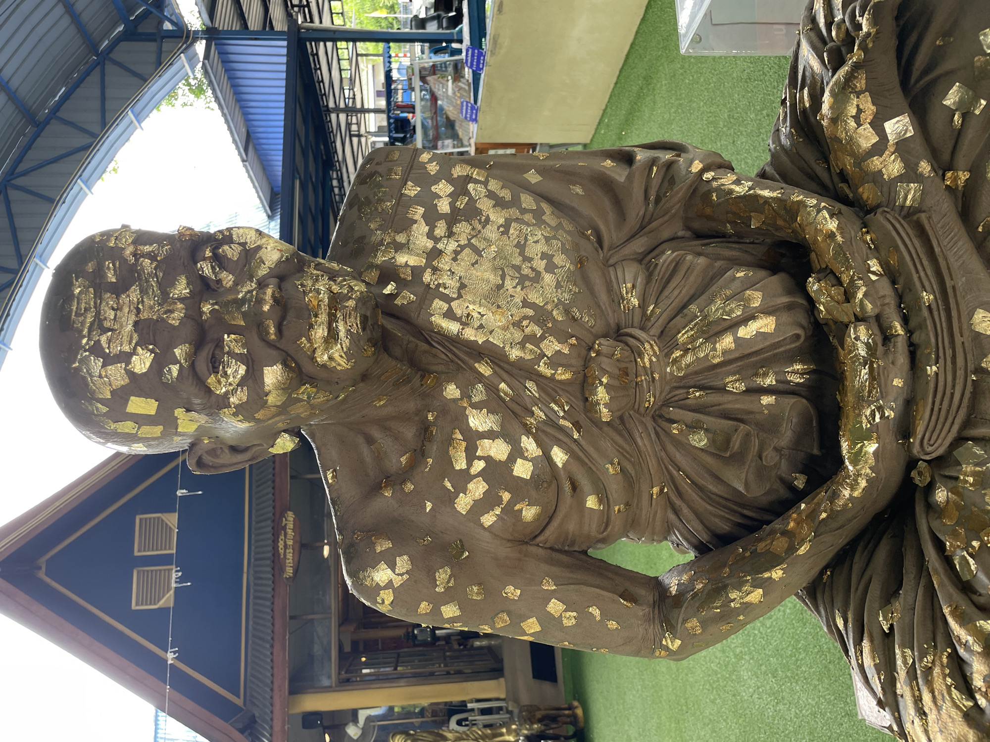 The image shows a statue of a Buddhist monk adorned with gold leaf, seated in a meditation pose. The statue is placed outdoors on a bright green artificial turf surface. In the background, there is a building with a dark blue triangular roof and a dark red trim. Part of the building features a sign with Thai script. The statue is the primary subject, covered in numerous squares of shimmering gold leaf, particularly on its face, robes, and hands. The lighting suggests it is daytime, with bright sunlight casting shadows. The overall environment appears to be a religious or cultural site in Bang Sare, Thailand.