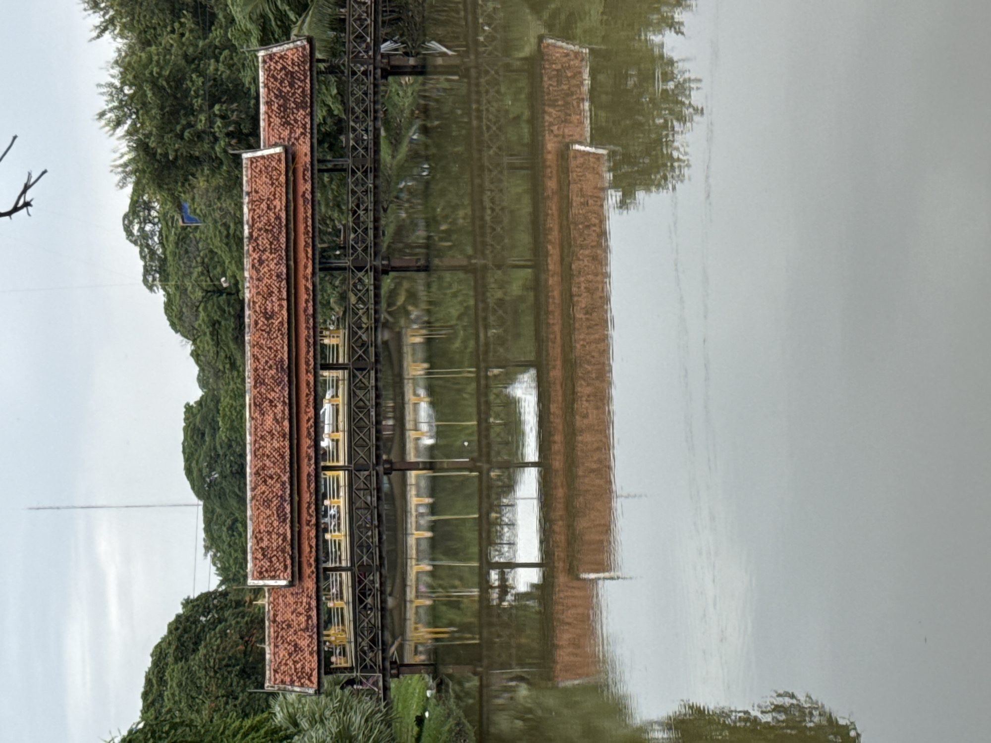 The image depicts a covered wooden bridge with a red tiled roof spanning over a calm body of water, likely a moat or canal, in Siem Reap, Cambodia. The bridge, constructed with dark wooden beams and featuring an ornate railing with yellow accents, reflects clearly in the water below. The bridge is lined with trees and foliage, with a tall antenna visible in the background, suggesting an urban or semi-urban setting. The sky is overcast, casting a soft light on the scene. There are a few indistinct figures visible on the bridge.