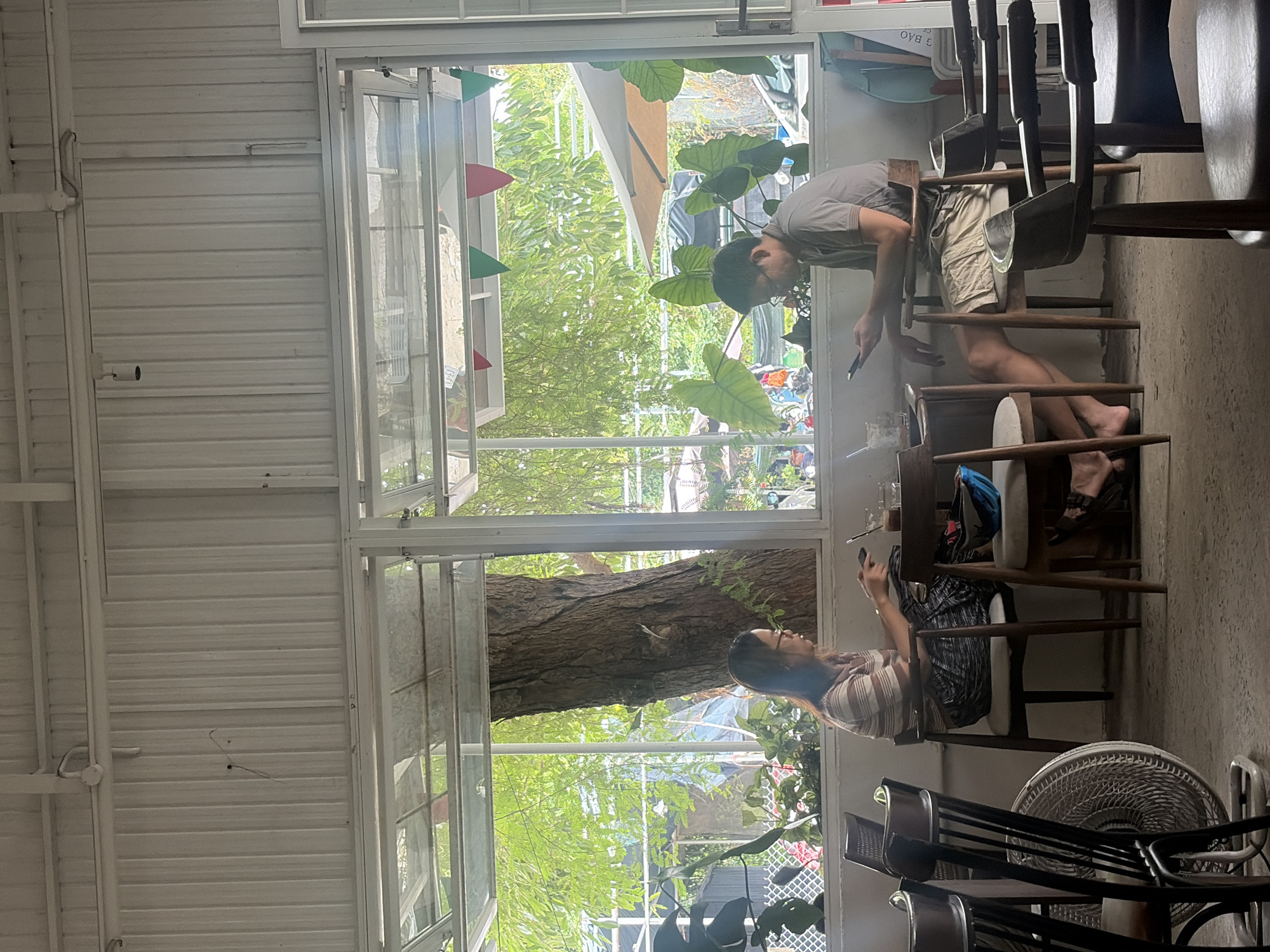 Two individuals, a woman and a man, are seated at separate tables in an outdoor cafe setting in Đà Nẵng, Vietnam. The woman, wearing glasses and a striped shirt, sits at a table with a drink and appears to be interacting with her phone. The man, dressed in a grey t-shirt and shorts, is also seated and focused on his phone. The cafe features wooden chairs and tables. Large windows overlook a lush green outdoor area with trees and plants. The ceiling is constructed of white corrugated metal. The overall atmosphere suggests a relaxed environment, possibly a coffee shop or restaurant.