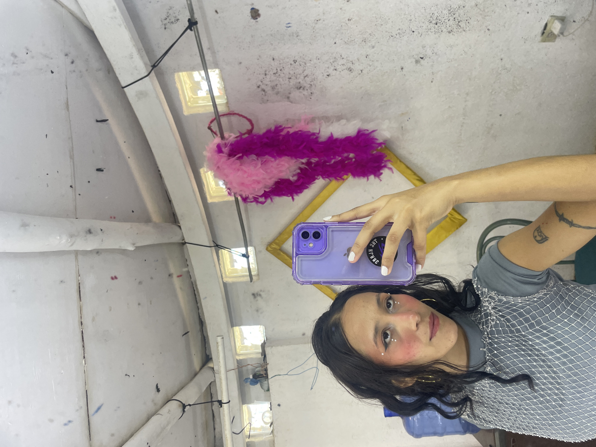 The image shows a young woman taking a selfie in what appears to be a rustic indoor space. The environment includes a concrete or plaster wall, a rough ceiling with exposed pipes and wiring, and small, barred windows. A pink and purple feather boa is hanging from a bar across the wall. The woman has dark, wavy hair, is wearing a grey top with a net overlay, and has glittery makeup around her eyes. She is holding a purple phone, which is capturing her reflection. A large blue water jug is visible in the background. There is a framed picture or object leaning against the wall to the right. The overall impression is of an informal, perhaps slightly bohemian or artistic, setting.