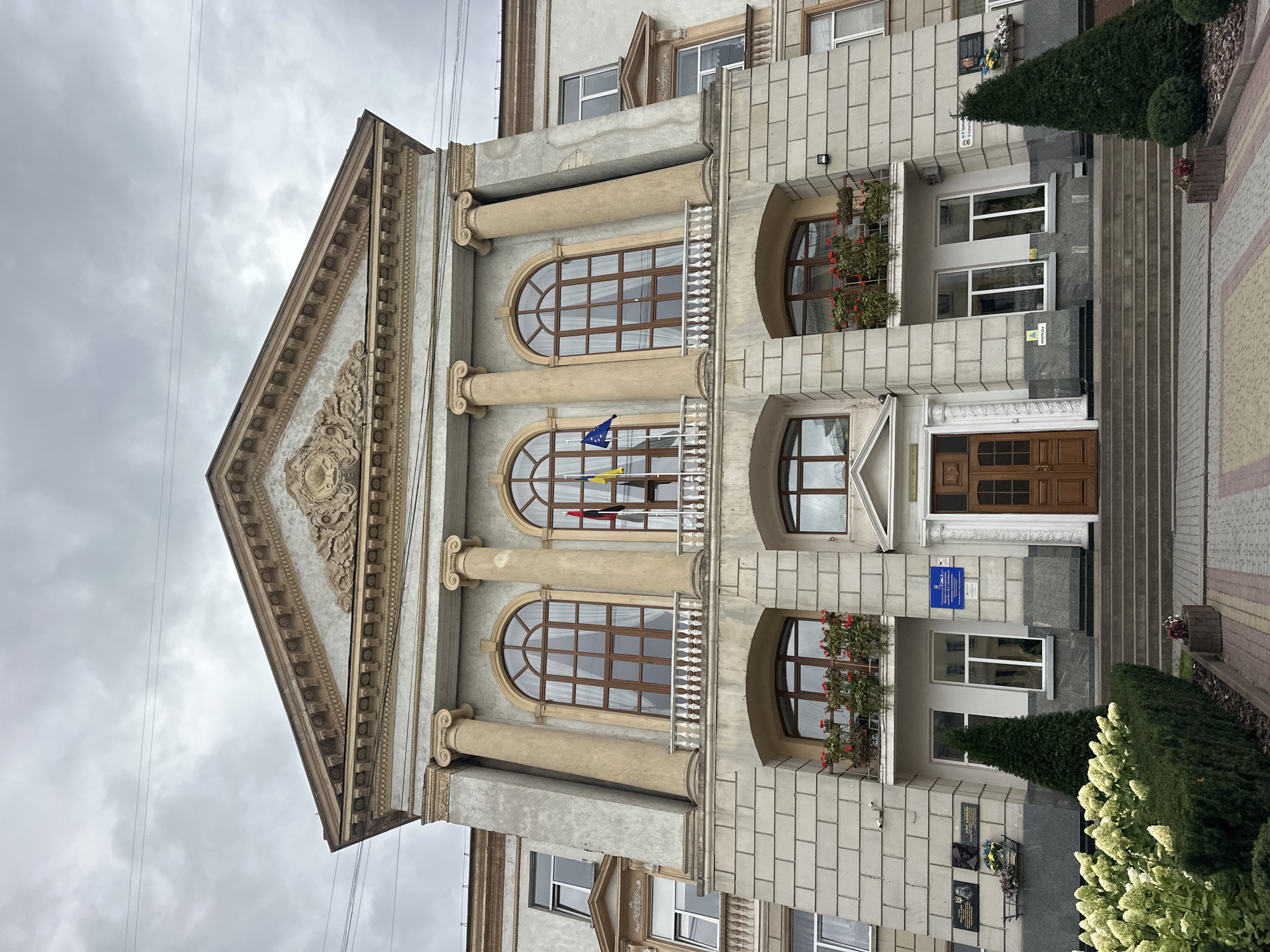 The image shows the facade of a building, which is the Ternopil State Technical University (TDTU). The building is made of stone and has a classical architectural style with columns, arches, and a pediment. There are several windows on the facade, some of which are arched and have decorative balconies. Flags are visible on the facade, suggesting an official or ceremonial occasion. The building is surrounded by greenery, including trees and bushes. The sky is overcast. The image appears to be taken from a low angle, looking up at the building.