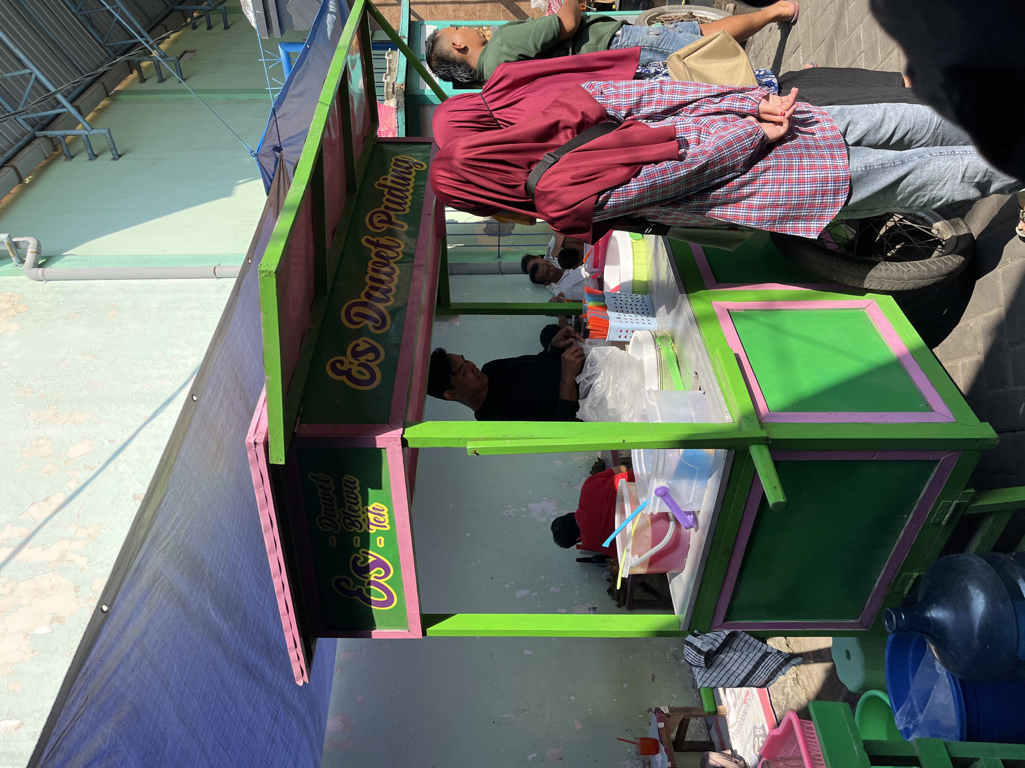An outdoor, medium shot captures a food cart selling "Es Dawet Puding" under a blue tarp. The cart is painted green with pink trim and features signage indicating its offerings. A vendor, a man in a black shirt, is preparing drinks inside the cart. Two women wearing maroon hijabs and plaid shirts are standing in front of the cart, facing away from the camera. Another man in a green shirt is visible to the right, and several other figures are present in the background. The environment appears to be a street or market area, with a textured, light-colored wall behind the cart and paving stones on the ground. The scene is illuminated by bright sunlight, casting shadows. The cart is equipped with various containers, cups, and straws, suggesting active business. The overall impression is of a bustling daytime scene at a local food stall in Juwet Kenongo, Indonesia.