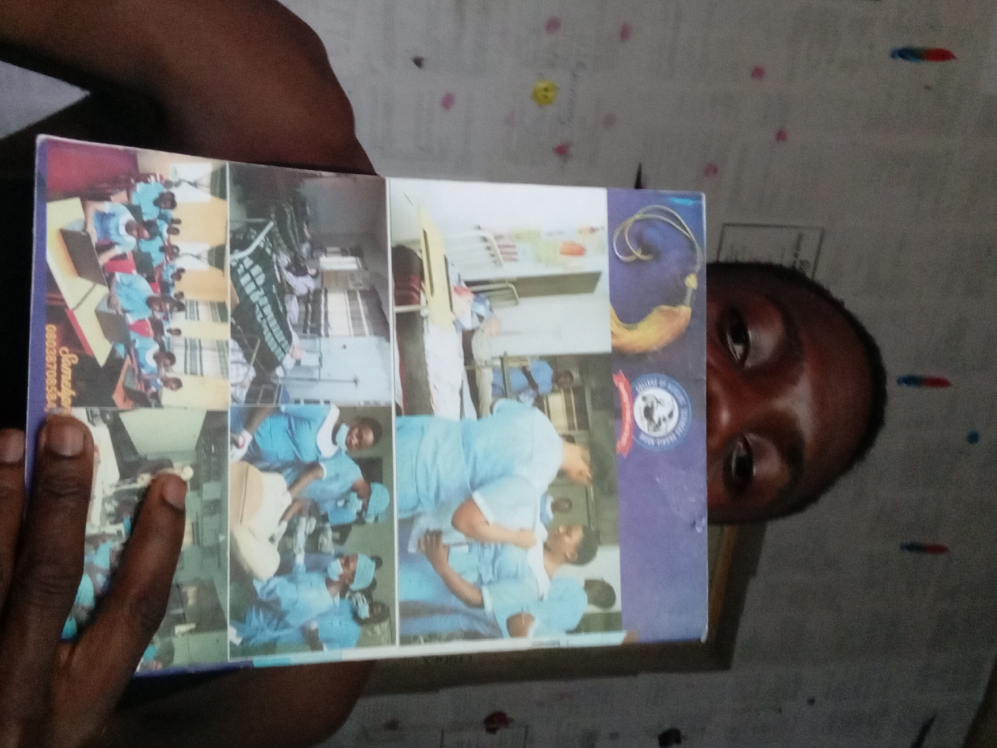A medium indoor shot shows a man holding a book open to display a collage of images to the camera. The collage predominantly features people in blue scrubs, suggesting a medical setting or training environment. Some images show them working at desks and in operating rooms. The book has a phone number printed on it: 08033970856. The man is partially visible behind the book, with his face partially covered by the book. The background is a white wall with some colorful marks or stickers. The scene suggests the man is sharing a visual record of an event or program related to medicine or healthcare, potentially in Akwete, Nigeria.