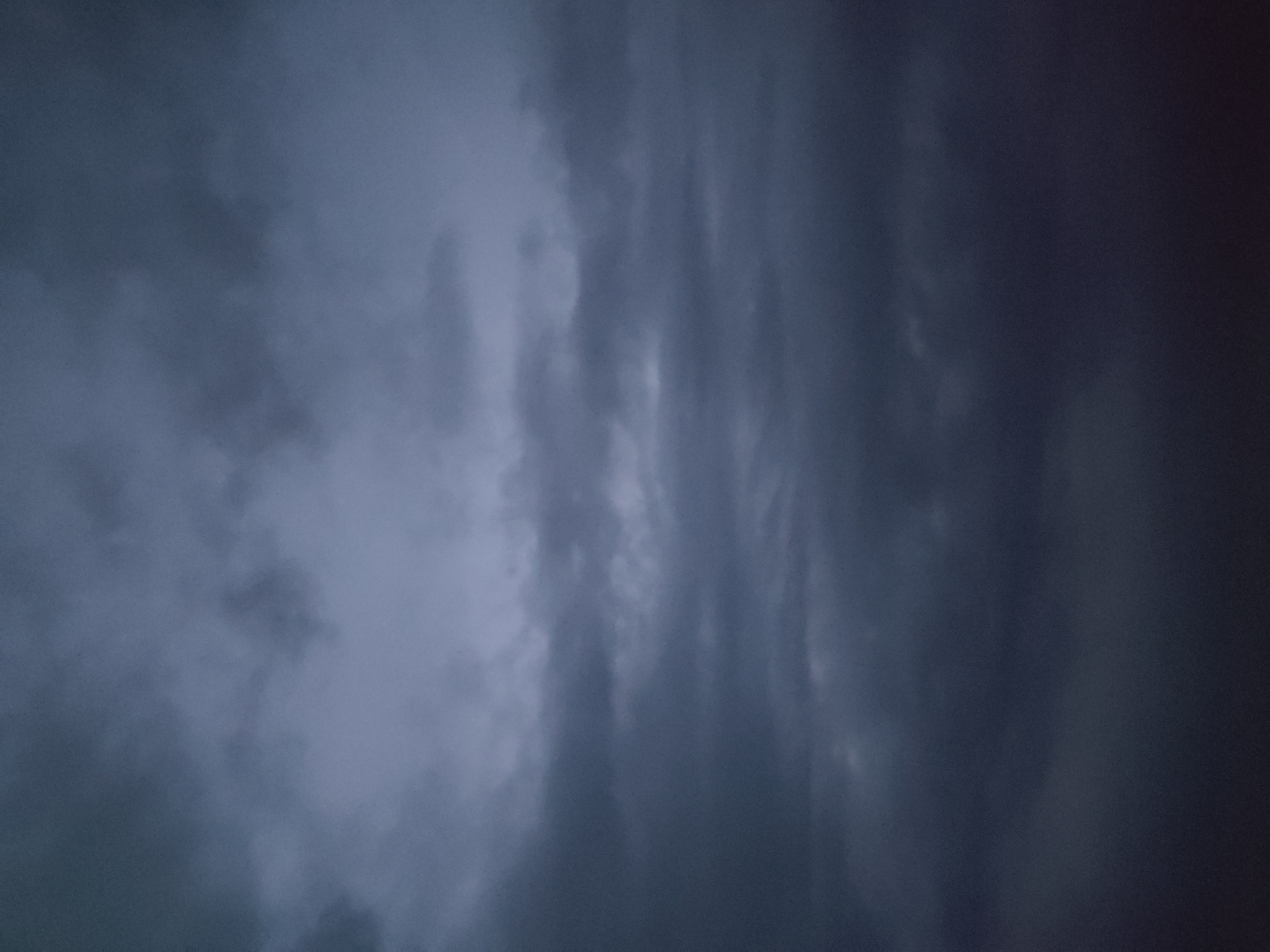 The image is a close-up, slightly blurry shot of dark, dramatic clouds. The clouds are predominantly shades of gray and deep blue, suggesting a stormy or overcast sky. There are variations in texture, with some areas appearing wispy and diffused, while others are denser and more defined, forming vertical streaks and swirling patterns. The lighting is dim, consistent with twilight or a heavily clouded day, and there are no distinct light sources visible. The composition is abstract, focusing solely on the cloud formations without any discernible horizon, land, or man-made objects. No people are visible. The overall mood conveyed is one of foreboding or powerful natural intensity. The image appears to be taken from an upward angle, looking directly into the sky. There is no visible text.