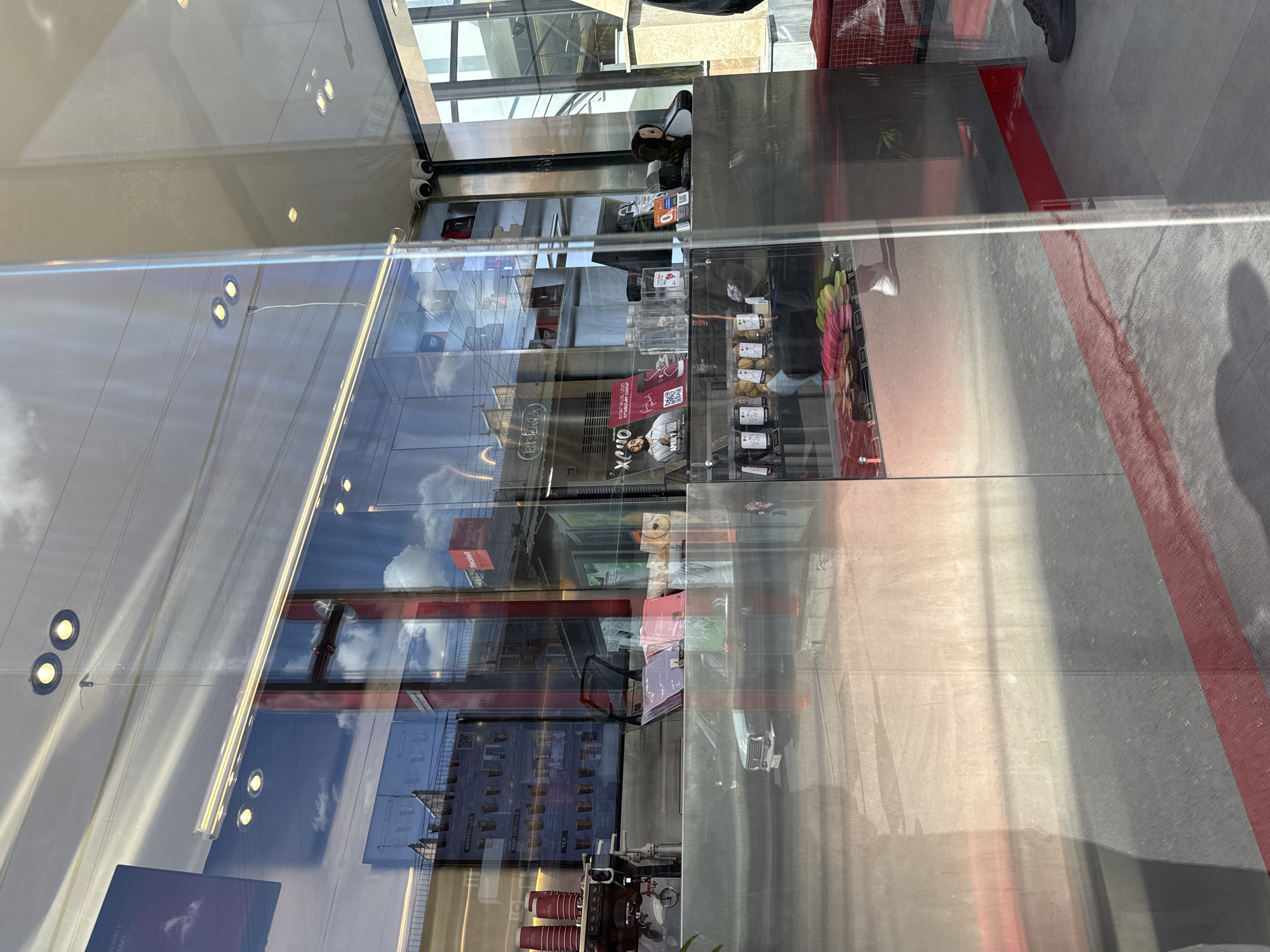The image, taken in Yerevan, Armenia, is a view through a glass storefront. The reflection shows a building across the street, the sky, and architectural details of the building where the photographer is standing. Inside the store, shelves are stocked with products including coffee beans and macaroons, arranged for sale. A coffee machine is visible on the counter. A person is standing on the outside sidewalk.