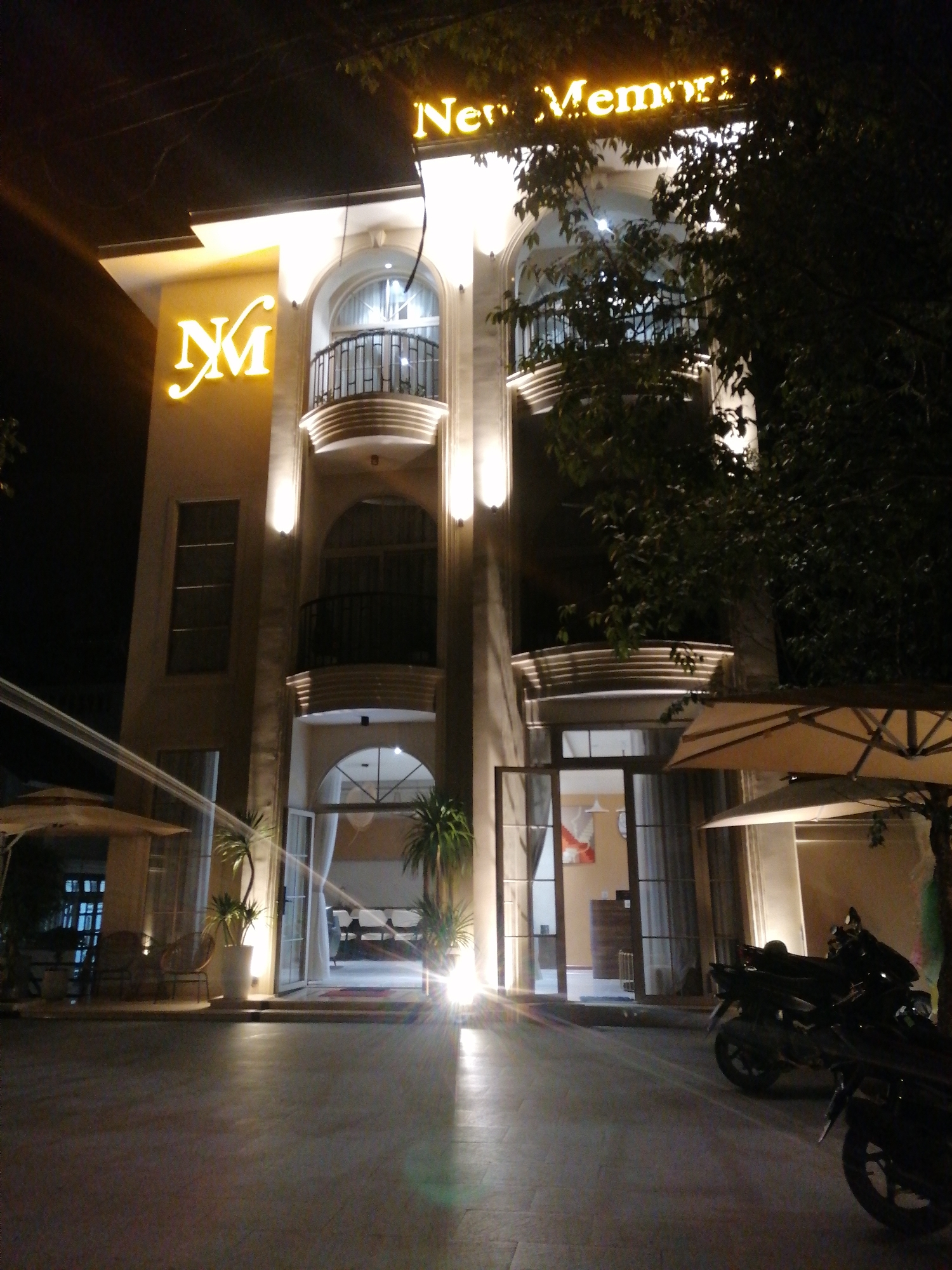 This is a night-time, eye-level, medium-shot photograph of the entrance of a hotel or establishment in Da Lat, Vietnam, identified by the neon signage which reads "New Memorial." The building features a modern facade with arched windows and balconies, illuminated by warm interior lighting and spotlights on the exterior. The main entrance is an open archway leading into a well-lit lobby area. In the foreground, the ground is paved with stone tiles and reflects the lights, creating a shimmering effect. Two motorcycles are parked to the right of the entrance. To the left, two outdoor umbrellas provide shade, and seating areas with chairs and tables are visible. The environment appears to be a well-maintained outdoor space, possibly a courtyard or garden area associated with the establishment. The overall impression is one of a welcoming and illuminated nighttime scene.