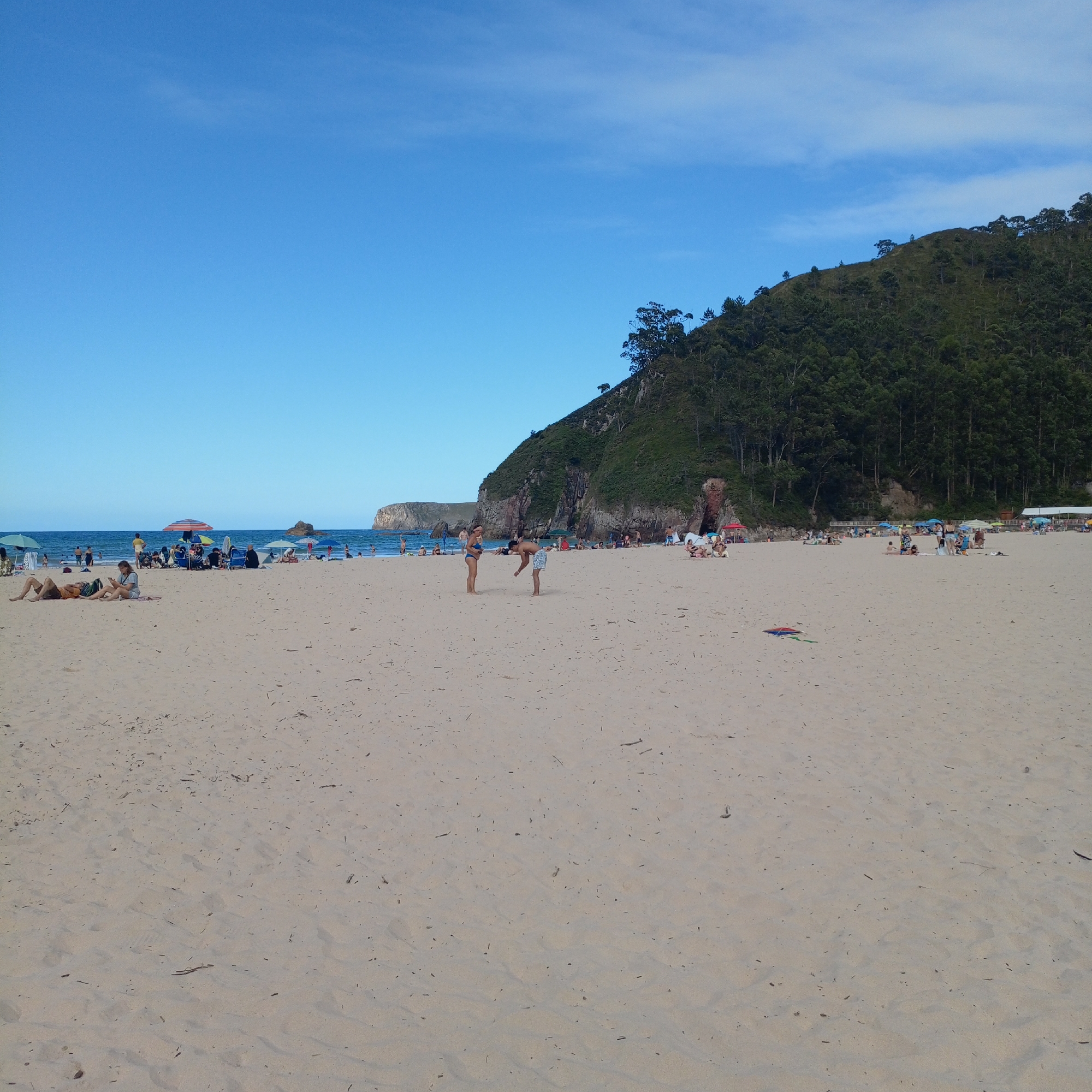 This is a medium-shot, horizontal, eye-level photograph of a crowded beach on a sunny day. The foreground is dominated by a sandy beach with some scattered debris. In the midground, numerous people are visible, some sitting under umbrellas, others walking or standing. Several colorful beach umbrellas provide shade. The ocean stretches out in the background, with gentle waves. On the right side of the image, a lush green, tree-covered hill rises steeply from the beach, with some rocky outcrops visible. The sky is a clear, vibrant blue. The scene suggests a warm, pleasant day at the beach, likely in the afternoon given the bright sunlight. The overall atmosphere appears relaxed and recreational, with people enjoying leisure time by the sea. There are no visible signs or text in the image.