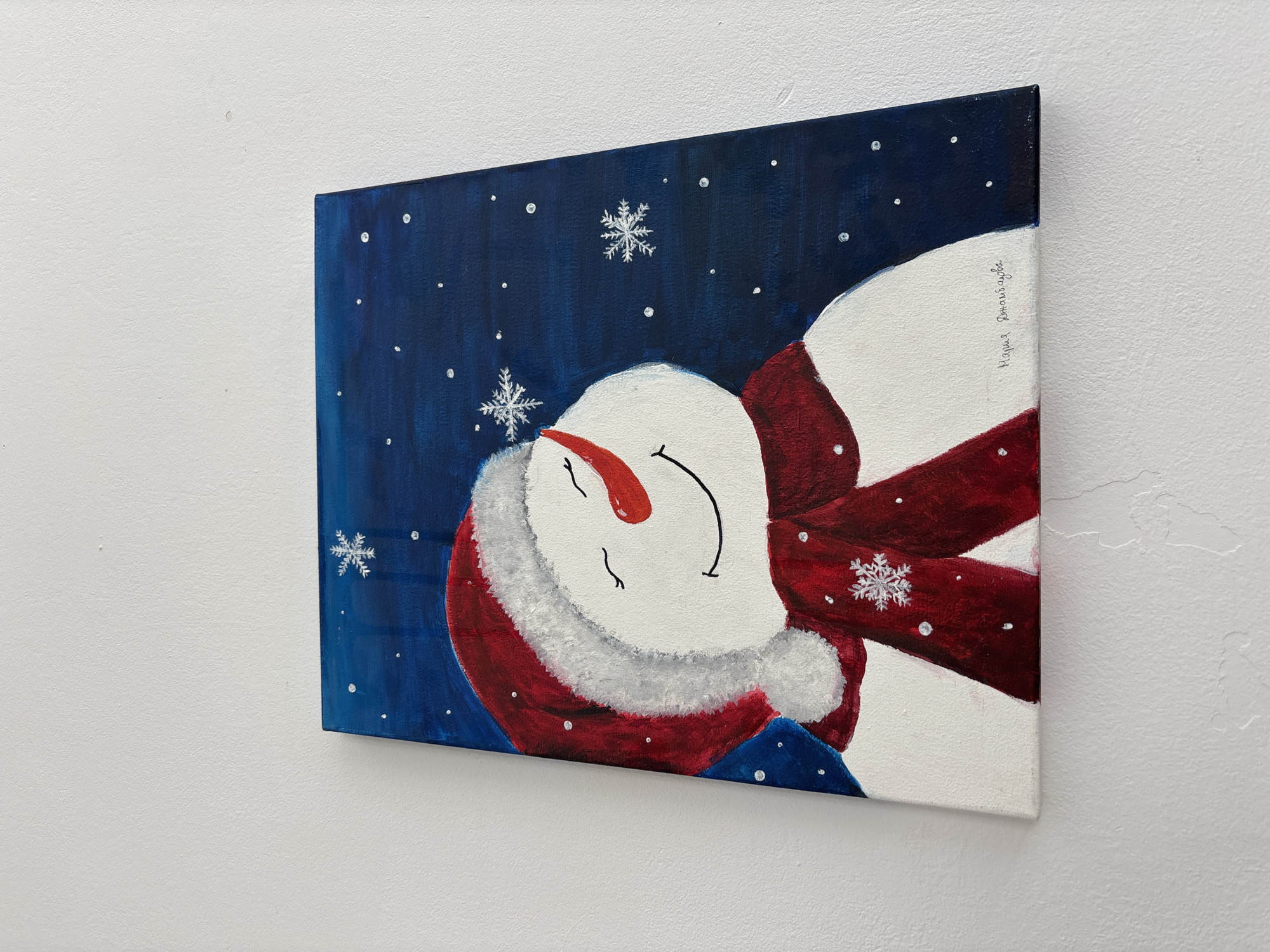An angled shot captures a vibrant painting on canvas, depicting a cheerful snowman against a deep blue background. The snowman, oriented diagonally, features a red hat with a white fluffy trim, a bright orange carrot nose, and a wide, curved smile with closed eyes, conveying a sense of peace or sleep. It wears a matching red scarf, also with a white snowflake detail.
The background is a dark blue night sky, dotted with numerous small white specks and several larger, detailed white snowflakes, suggesting a winter wonderland. The painting exhibits rich primary colors: deep blue, bright white, and vibrant red. The canvas is mounted on a subtly textured white wall, seen in the surrounding areas of the image.
Vertical text, likely a signature, is visible on the right side of the snowman's white body, written in what appears to be Cyrillic: "Мария Домницова". The artwork evokes a festive and serene winter holiday spirit. This scene is located in Zlatograd, Bulgaria.
