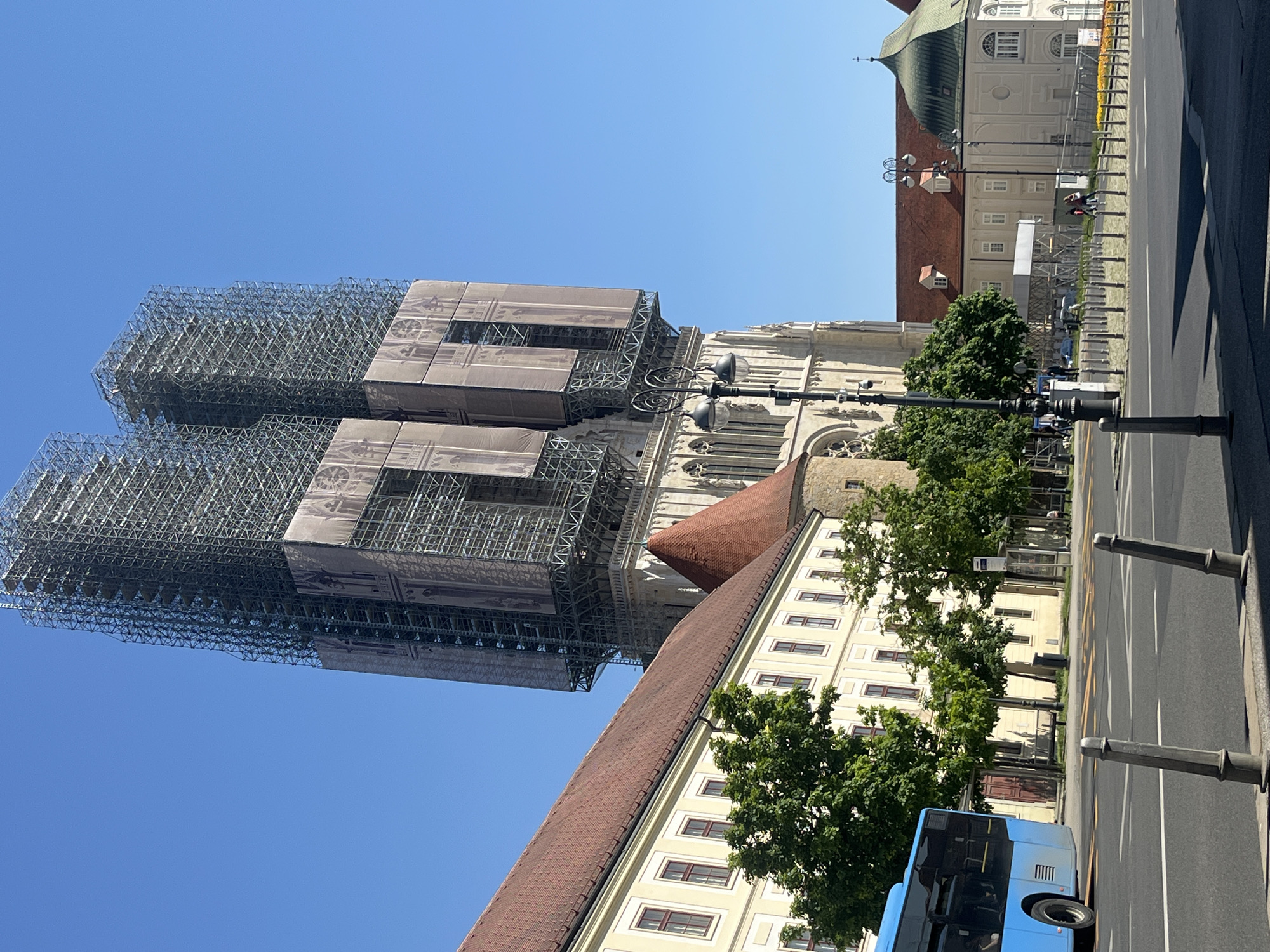 The image shows the Zagreb Cathedral in Croatia, with its two towers undergoing significant construction. The towers are enveloped in extensive scaffolding, partially covered with printed scrims that visually suggest the appearance of the towers underneath.
Next to the cathedral is another building with a red tiled roof, the facade colored in shades of yellow and beige.
The view is taken from street level. The street itself is paved and marked with painted lines. A modern blue bus is partially visible in the lower right corner of the frame. Trees line the street, offering a green contrast to the surrounding architecture. A street lamp stands in front of the cathedral. Beyond the cathedral are glimpses of other buildings. The sky is clear and blue.