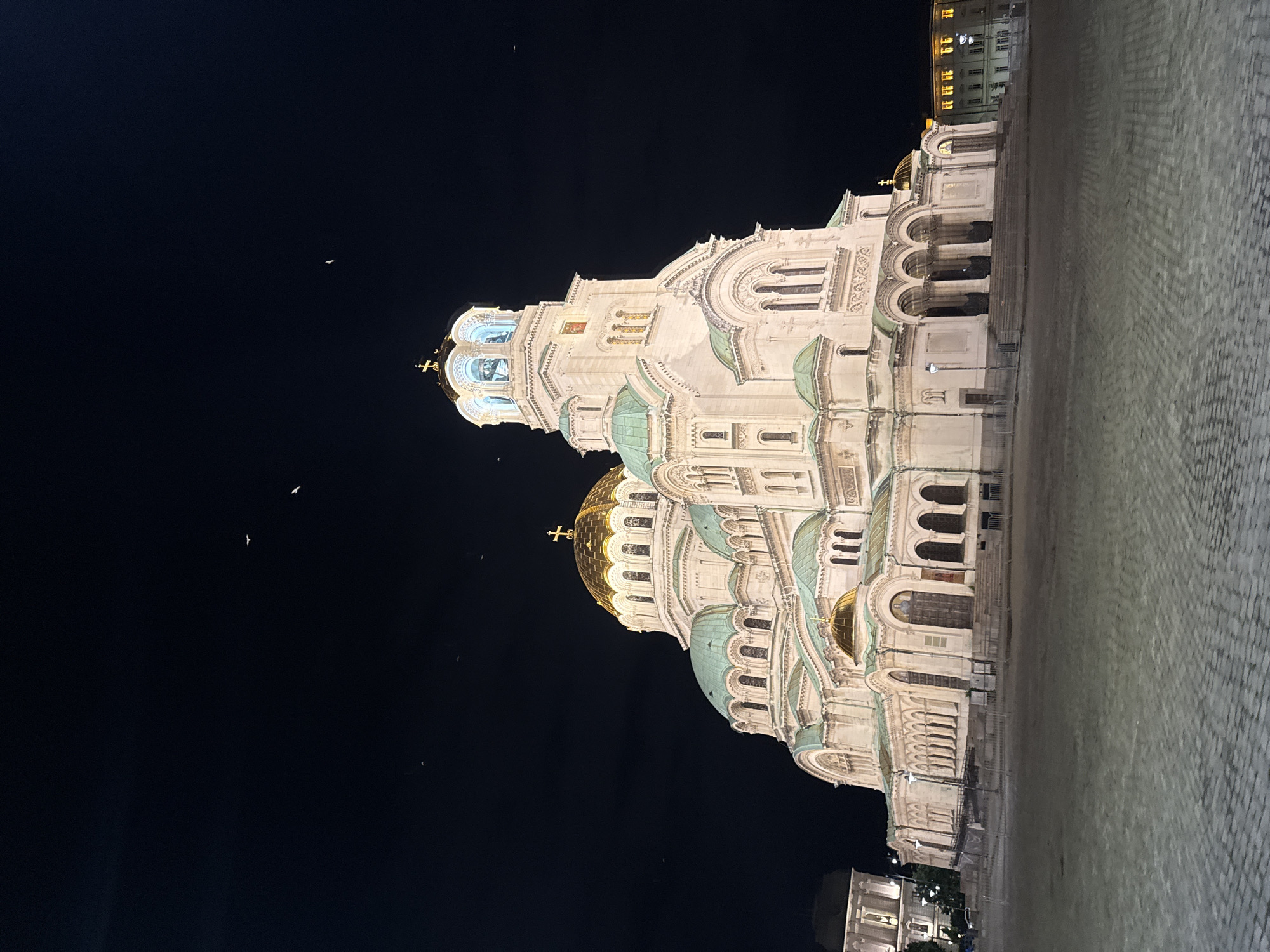 The image shows the Alexander Nevsky Cathedral in Sofia, Bulgaria, at night. The cathedral is a large, imposing structure with multiple domes, including a prominent golden dome and several green domes. The facade is illuminated, highlighting its intricate architectural details. A bell tower rises above the main structure, also lit. The sky above is dark and appears to have a few small birds flying in it. In the foreground, there is a cobblestone plaza. To the left and right of the cathedral, other buildings are visible, some also illuminated. The overall scene is one of a grand, well-lit religious monument against a dark night sky.