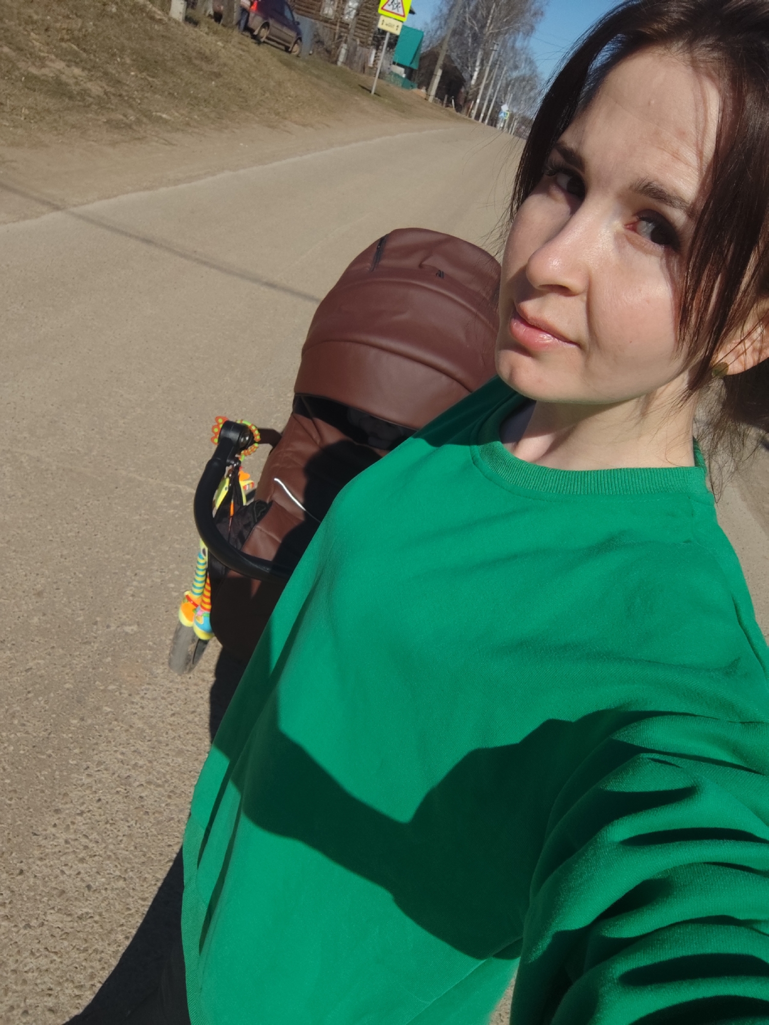 The image is a selfie of a woman and a baby stroller on a sunny day in what appears to be a rural or semi-rural setting in Фоки, Russia. The main subject is a woman with dark hair, looking towards the camera with a calm expression. She is wearing a vibrant green long-sleeved top.
To her left, a brown baby stroller is partially visible, with a colorful, multi-ringed toy attached to its handle. The woman is holding the camera to take the photo, positioning herself to the right side of the frame.
The background reveals a paved road under a clear blue sky. On the left side of the road, there is a grassy, slightly sloping bank. Further down the road, several bare deciduous trees and a few residential structures, likely houses, can be seen. A dark-colored car is also visible in the distance on the left.
Notable details include the strong, bright sunlight, which casts distinct shadows of the woman and the stroller across the road and her green top, indicating mid-morning or afternoon. A yellow diamond-shaped road sign, partially obscured, is visible on the left side of the road, suggesting a warning for crossroads or a dangerous intersection. The bare trees imply the season is either early spring or late autumn. The overall scene suggests a leisurely walk outdoors. No prominent visible text other than the partial road sign.