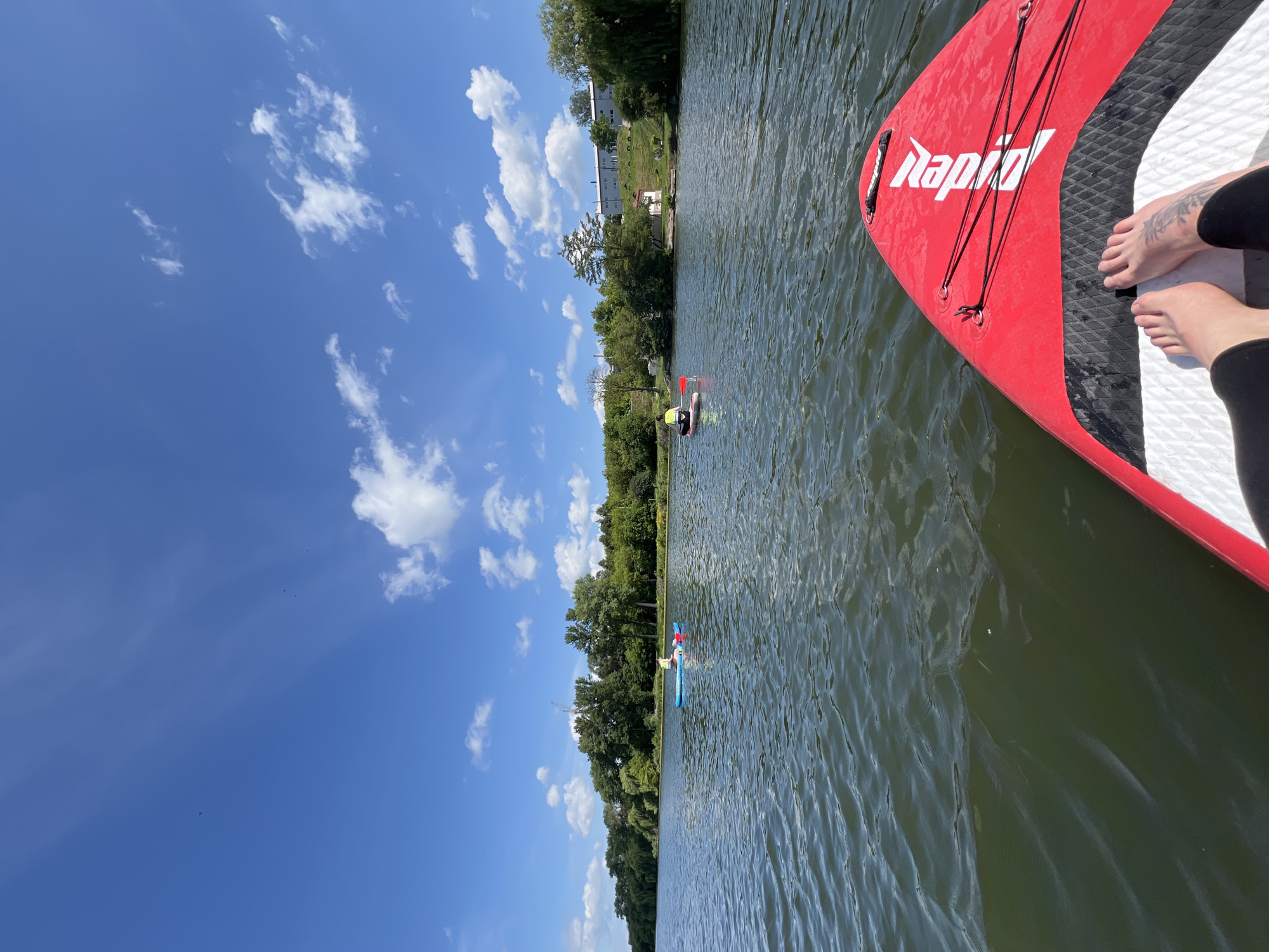 The image depicts a sunny day with a clear blue sky and scattered white clouds over a body of water. The foreground shows the front of a red paddleboard with the white logo "RAPID" visible. A person's bare feet are positioned on the paddleboard, with visible tattoos on the left ankle. Two other individuals are paddleboarding in the distance on blue and white paddleboards. The background features a tree-lined shore with buildings and a construction crane visible in the far distance. The location is specified as Bila Tserkva, Ukraine.