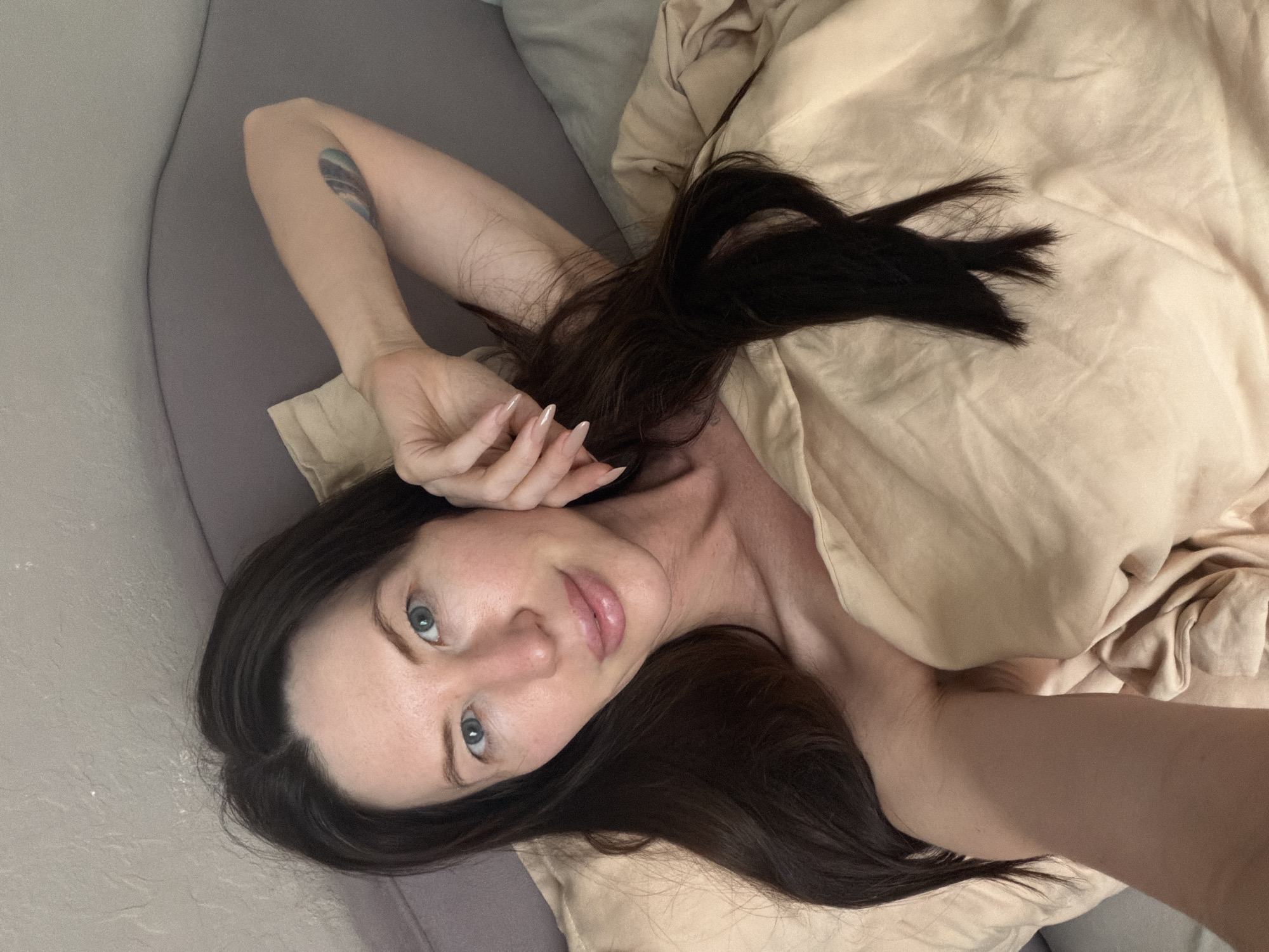 The image captures a woman lying on a bed indoors. She is positioned diagonally across the frame, with her head towards the bottom left and her body extending towards the upper right. Her long, dark hair is spread out on the pillow and blanket. She is partially covered by a light beige or tan duvet/blanket, with her bare shoulder and upper chest visible.
The woman has light skin, striking light blue or green eyes, and full lips. Her expression is calm and direct, looking towards the camera. One arm is extended above her head, with her hand resting near her face. Her nails are long and manicured. On her left upper arm, a distinct tattoo is visible, appearing to be an abstract or naturalistic design, possibly depicting a wave or landscape in muted blues and greens.
The bedding consists of a light grey fitted sheet or mattress cover on one side, and a beige/tan pillow and duvet/blanket. The setting appears to be a bedroom, suggesting a moment of relaxation or just waking up. The lighting is soft and even, typical of indoor ambient light, making it difficult to determine the exact time of day, though it suggests daytime. There is no visible text in the image. The image is set in Perm, Russia.