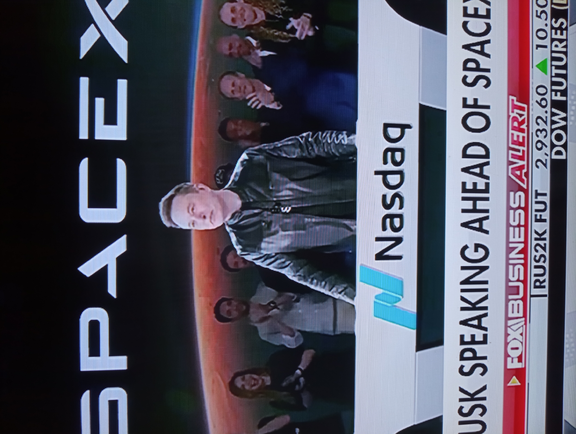 The image shows a media broadcast featuring Elon Musk in front of a "SPACEX" logo, with a view of a red planet behind him. Below this, a Nasdaq logo is visible, and text reads "MUSK SPEAKING AHEAD OF SPACEX". A Fox Business Alert banner is also present, along with stock market data for "RUS2K FUT" and "DOW FUTURES". The scene depicts a formal event or announcement, likely related to SpaceX's business activities. The image quality suggests it was captured from a screen, possibly a television or computer monitor. There is no indication of the location being the city of Mahilyow, Belarus; the context points towards a financial or technological hub.