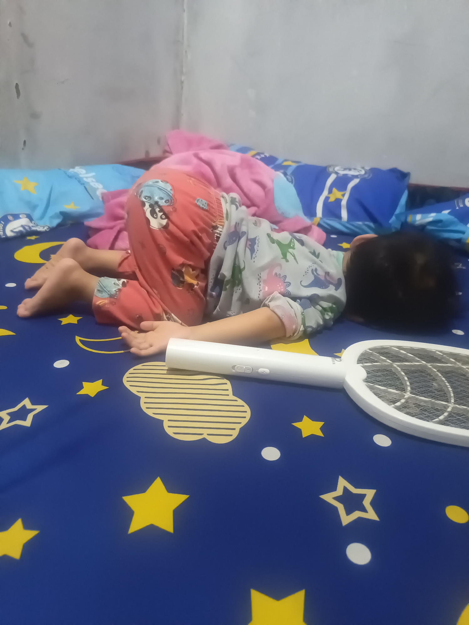A child is lying face down on a blue mattress with yellow stars and white circles. The child is wearing red pajamas with a panda pattern and a light-colored shirt with a dinosaur pattern. A white electric mosquito swatter is on the mattress near the child. The scene is likely indoors, possibly in a bedroom, in the city of Karangkedawung, Indonesia.