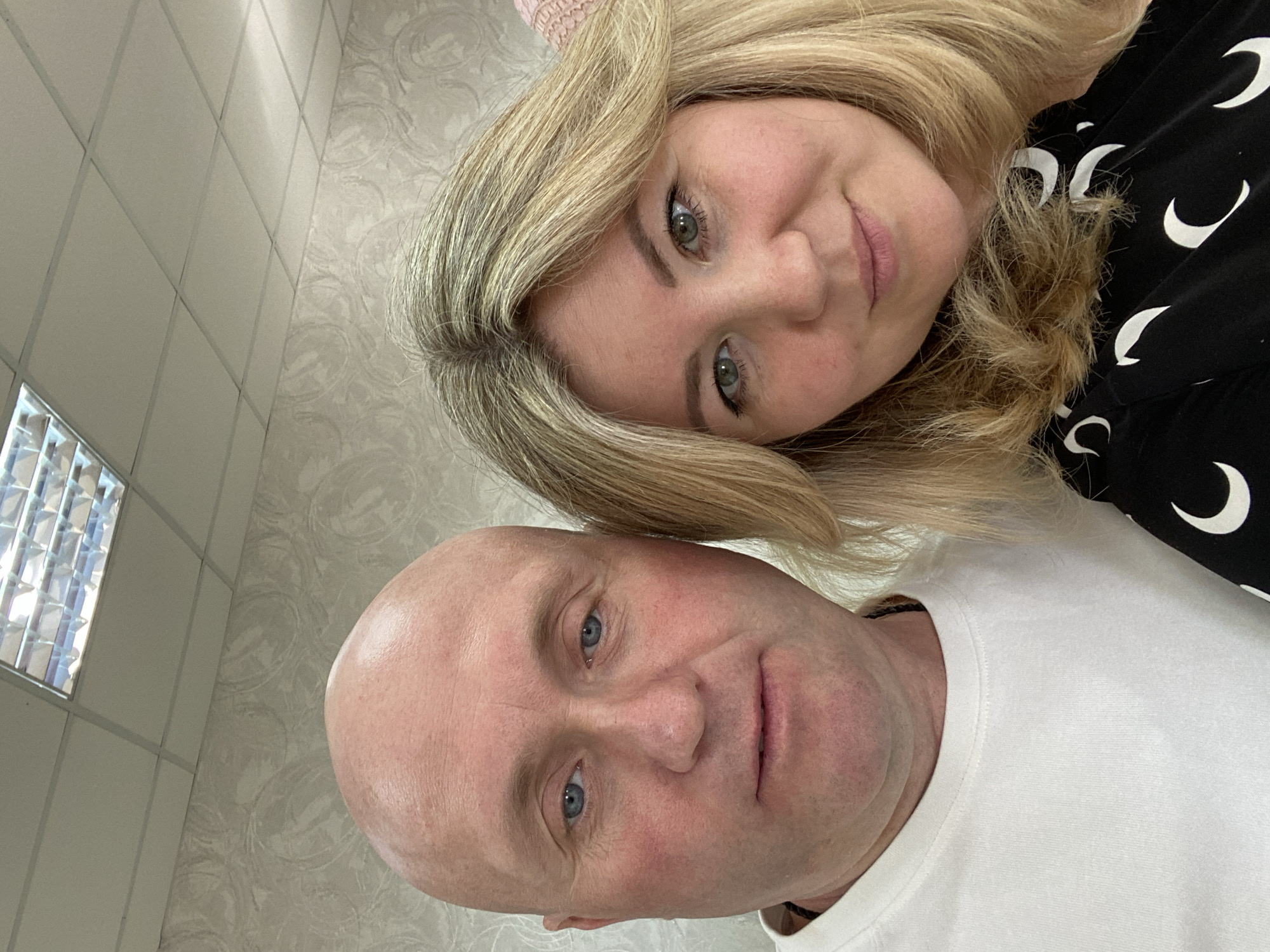 The image is a close-up selfie of two people, a bald man and a woman with blonde hair, positioned in front of a wall with a patterned wallpaper and a tiled ceiling with fluorescent lighting. The man, on the left, is wearing a white t-shirt and has blue eyes and fair skin with some redness on his cheeks. The woman, on the right, has green eyes, blonde hair, and is wearing a black top with a crescent moon pattern. They are both looking towards the camera. The scene appears to be indoors, possibly in a home or office environment. The location is not explicitly identifiable as Chaikovsky, Russia, based solely on the visual information provided.
