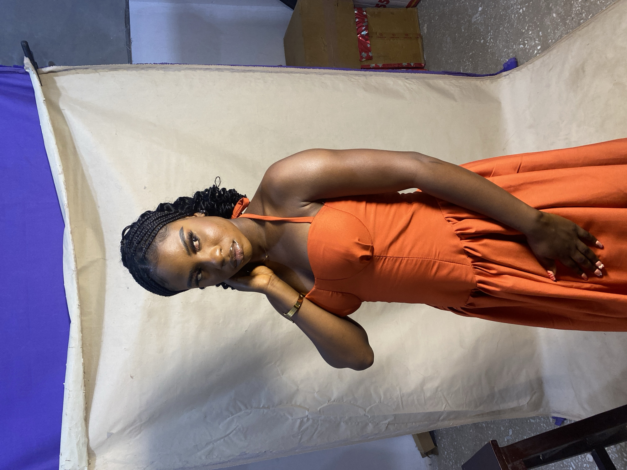 A young, dark-skinned woman is photographed in a studio setting in Jos, Nigeria. She is wearing an orange dress and has braided hair. Her left hand rests on her face, and her right hand is placed on her abdomen. She is wearing a gold bracelet on her left wrist. The background consists of a makeshift studio setup with a cream-colored sheet draped over what appears to be a purple-colored surface. Boxes and a chair are visible in the background. The lighting is artificial, illuminating the woman and the backdrop.