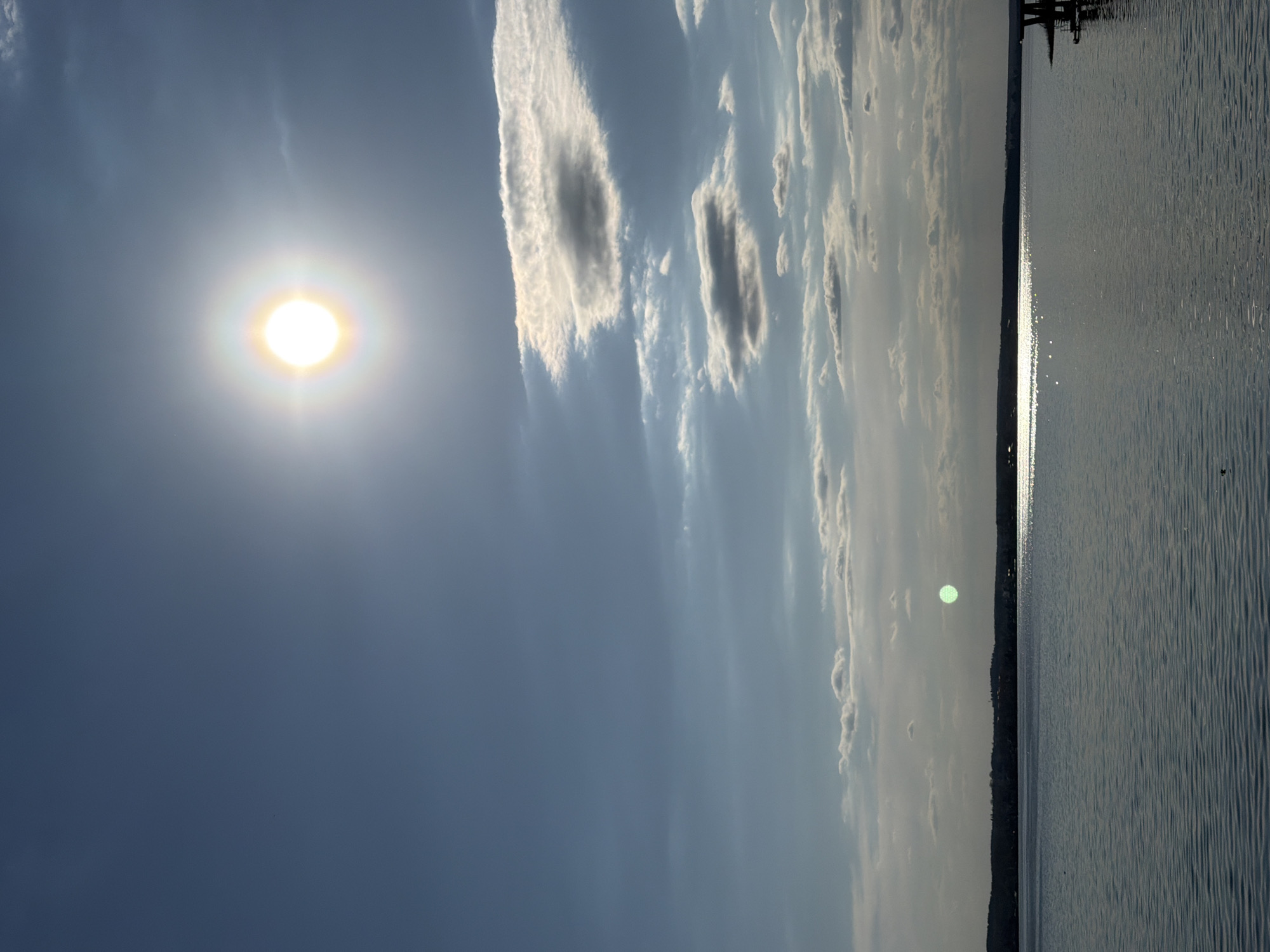The image depicts a bright, sunny day over water, possibly a lake or bay. The sun is prominent in the upper left quadrant of the image, surrounded by a blue sky with scattered clouds. A narrow strip of land, possibly a shoreline, is visible along the right edge of the image, separating the sky from the water. There are ripples on the water's surface. At the upper edge is a small structure, possibly a dock. Given the request for the location to be in Votkinsk, Russia, it is possible this image depicts a scene on the Votkinsk Reservoir. There are no people visible in the image.