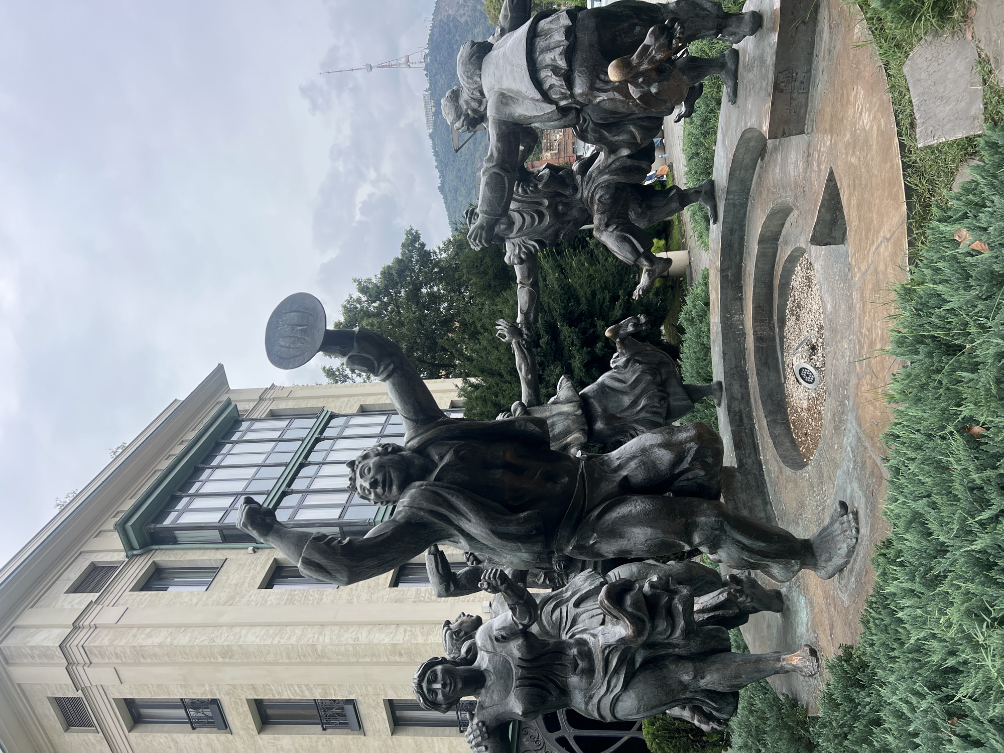 This image depicts the "Chronicle of Georgia" monument, also known as the Tbilisi Sea monument, located on a hill overlooking Tbilisi, Georgia. The monument is a large, imposing structure featuring bronze sculptures of historical Georgian figures and scenes. In the foreground, a group of dynamic bronze figures are captured mid-motion, appearing to be dancing or celebrating. The central figure, a man, raises his arms and holds a round object, possibly a tambourine or shield. Other figures, both male and female, are depicted in various poses of movement, with flowing garments and expressive faces. They are arranged in a circular formation around a shallow fountain basin.
The monument stands against a backdrop of a building with a light-colored, textured facade and large windows. In the distance, lush green trees are visible, and further beyond, a television tower can be seen on a distant mountain, a prominent landmark of Tbilisi. The sky is overcast with clouds, suggesting a temperate weather day. The surrounding area is landscaped with dense, green shrubs. The overall scene conveys a sense of historical significance and vibrant cultural expression.