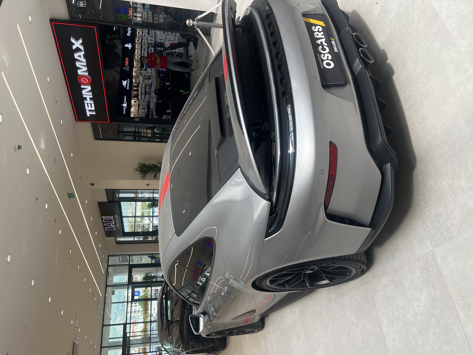 Here is a description of the image:
The image shows the rear of a gray Porsche GT3, likely on display inside a shopping mall. The car has a red stripe running along its roof. The angle is from a high vantage point, focusing on the rear details like the wing, exhaust pipes, and the "OSCARS" license plate.
In the background, the mall environment is visible with stores such as "TEHNOMAX" and "BABI". "TEHNOMAX" appears to be an electronics store, recognizable by the displays inside. "BABI" is visible in the background. The mall has a modern interior with bright lighting and glass storefronts.
Based on the text on the license plate ("OSMANAGIĆ CO") and the store names, the scene is located in Bjeliši, Montenegro.