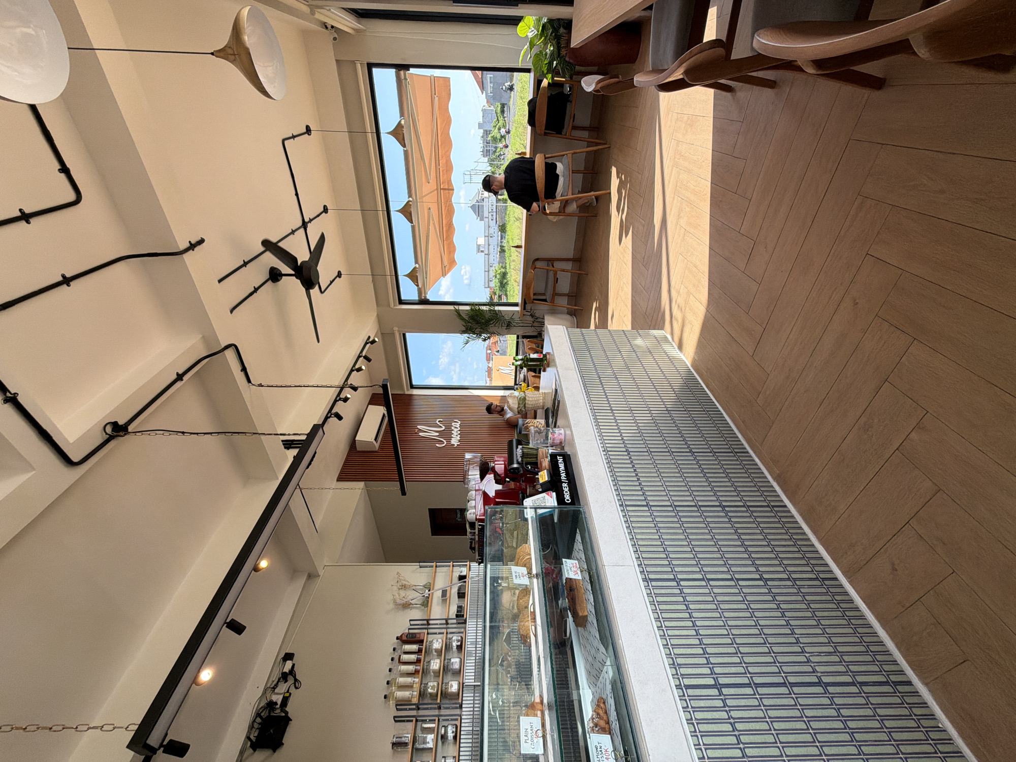 The image captures the interior of a cafe or restaurant, likely in Canggu, Indonesia, given the tropical landscape visible through the windows. The space features a long, glass-fronted display case filled with baked goods, suggesting it is a bakery or patisserie. A worker stands behind the counter, presumably preparing orders, while a customer sits at a table near the window, enjoying the view. The interior design includes overhead lighting fixtures and black piping against a white ceiling, contributing to a modern aesthetic. The flooring transitions from a patterned tile near the display case to wooden planks in the dining area. The overall atmosphere is bright and airy, enhanced by natural light streaming through the windows.