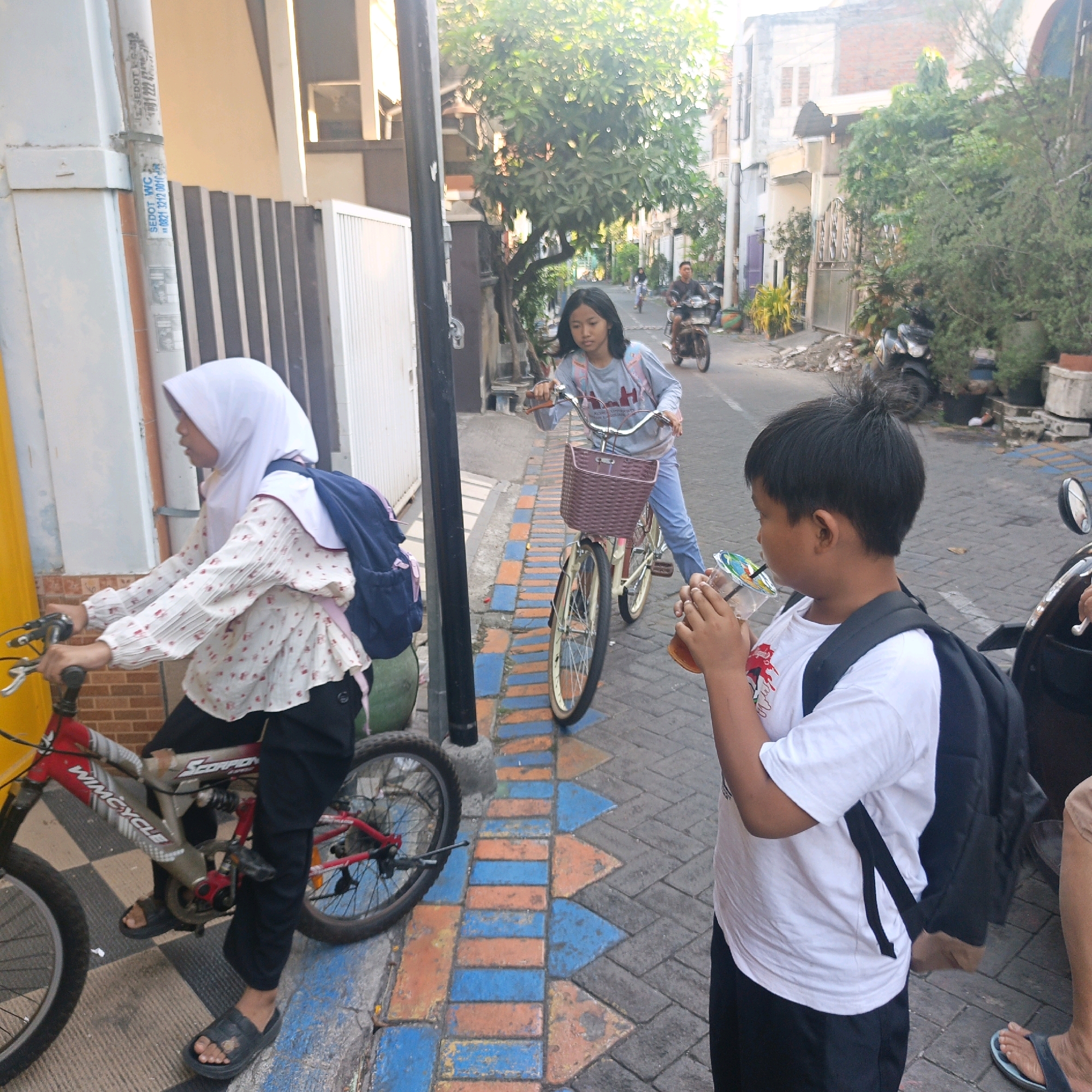The image depicts a street scene in Surabaya, Indonesia, with several individuals and bicycles. In the foreground, a young boy wearing a white t-shirt and black shorts, with a black backpack, is drinking from a clear plastic cup with a colorful lid. To his left, a girl wearing a white hijab and a patterned long-sleeved shirt with a navy blue backpack is seated on a red bicycle. Her feet are in black sandals. The bicycle has "WIMCYCLE" printed on the frame. Further down the street, another girl in a gray long-sleeved shirt and blue pants is riding a bicycle with a woven basket. In the background, a man is riding a motorcycle. The street is paved with interlocking bricks featuring a pattern of orange and blue rectangles and arrows. Buildings line the street, with trees and vegetation visible. A utility pole with signage is also present. The overall scene suggests a casual daytime activity, possibly commuting or recreation.