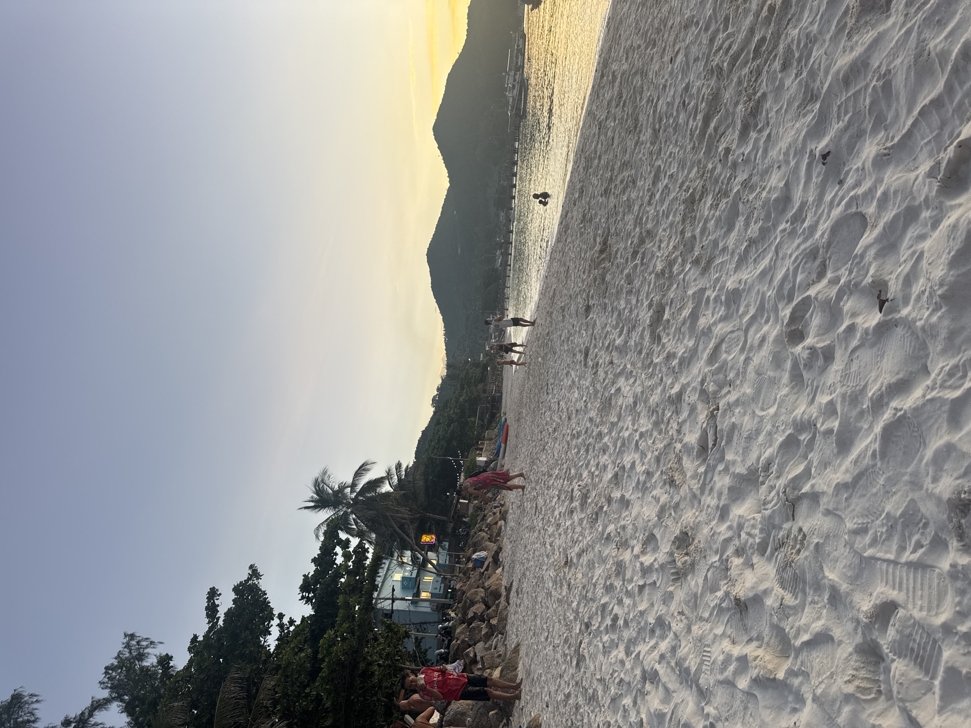 The image depicts a beach in Ko Pha Ngan, Thailand during the golden hour. The sand is white and textured with footprints. In the foreground, the beach stretches towards the sea. In the mid-ground, several people are visible on the sand and in the water. A few individuals are walking along the shoreline, and a couple are wading in the shallow water. Behind them, a line of rocky shore is dotted with trees and palm trees, leading to buildings, some of which are illuminated. Further in the distance, lush green hills form a backdrop against a pale yellow and blue sky. Boats are anchored in the calm sea. The overall scene conveys a peaceful and relaxed atmosphere.