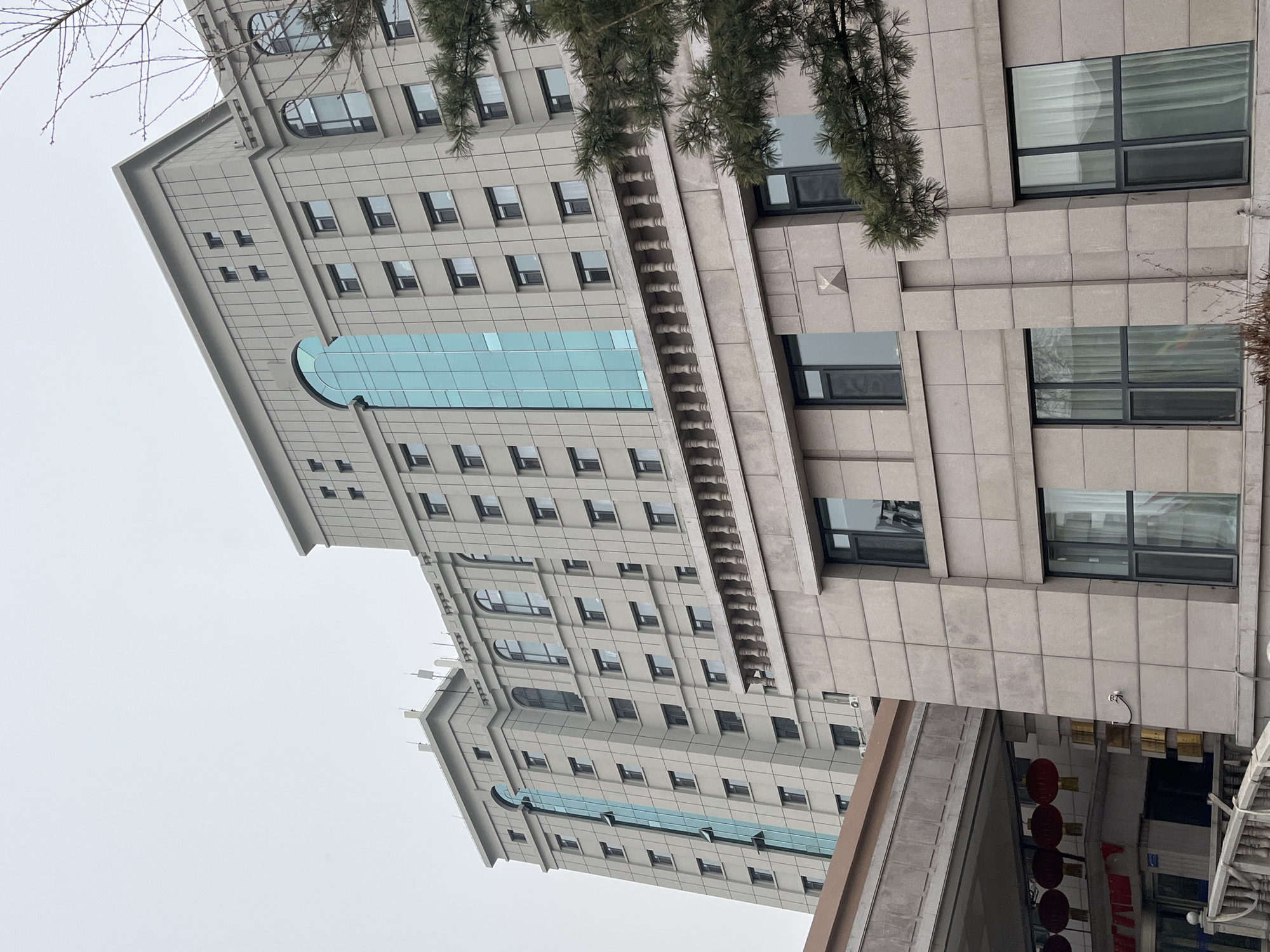 The image captures an upward-angled view of a multi-story building in Changchun City, China. The building features a gray facade with rectangular windows, with a central vertical section of blue glass. A tree branch is visible in the upper right, obscuring part of the building. Red Chinese lanterns are also visible in the lower right corner, hanging in front of a shop. The sky is overcast and gray.