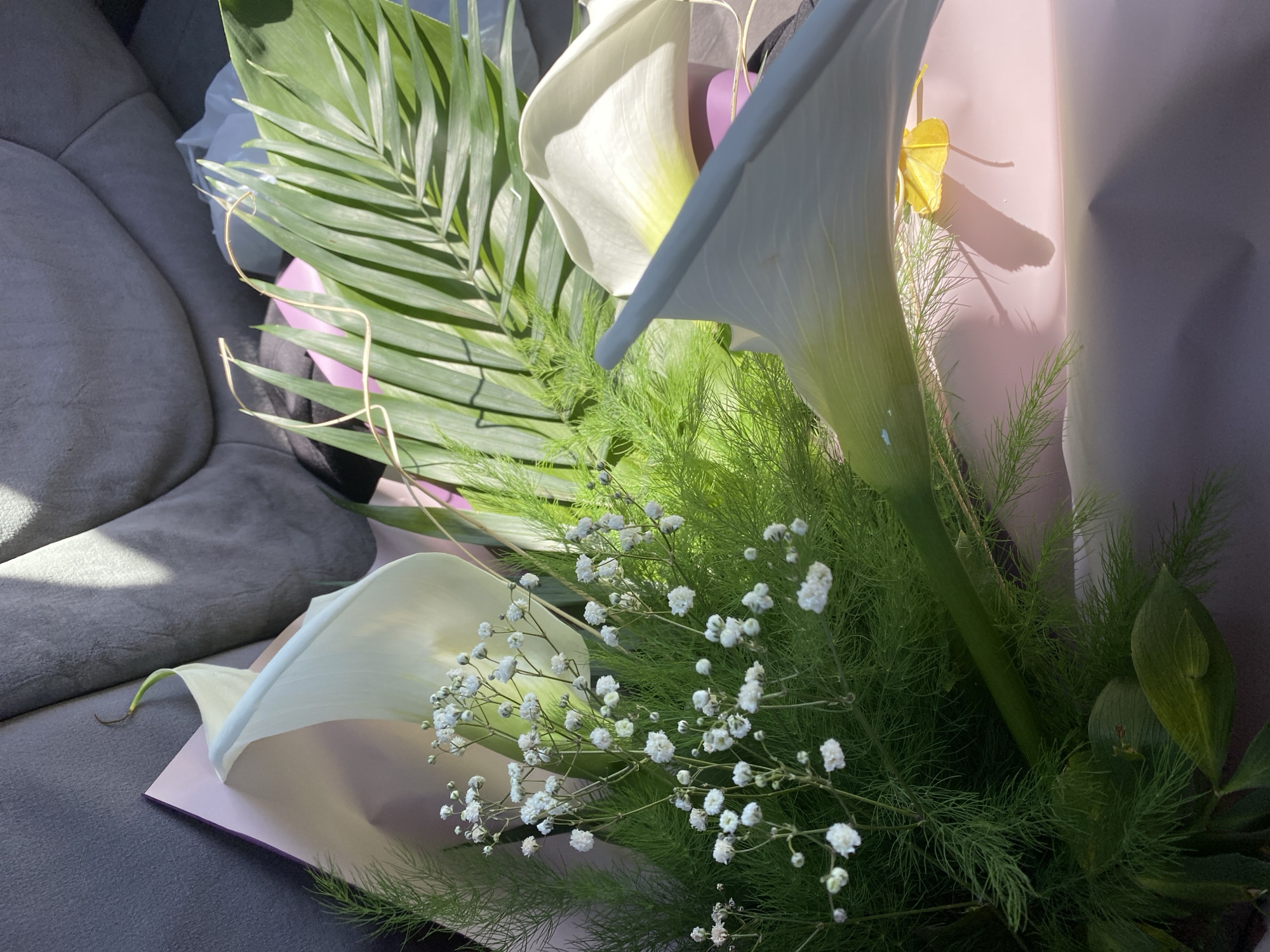 The image shows a bouquet of flowers, primarily composed of white calla lilies, baby's breath, and various green foliage, wrapped in light pink paper. The bouquet rests on a gray surface, possibly the seat of a car or a piece of furniture. The lighting suggests an indoor environment, possibly during the daytime. There are no people visible in the image. There is no information available in the image about the location being in the city of Varna, Bulgaria.