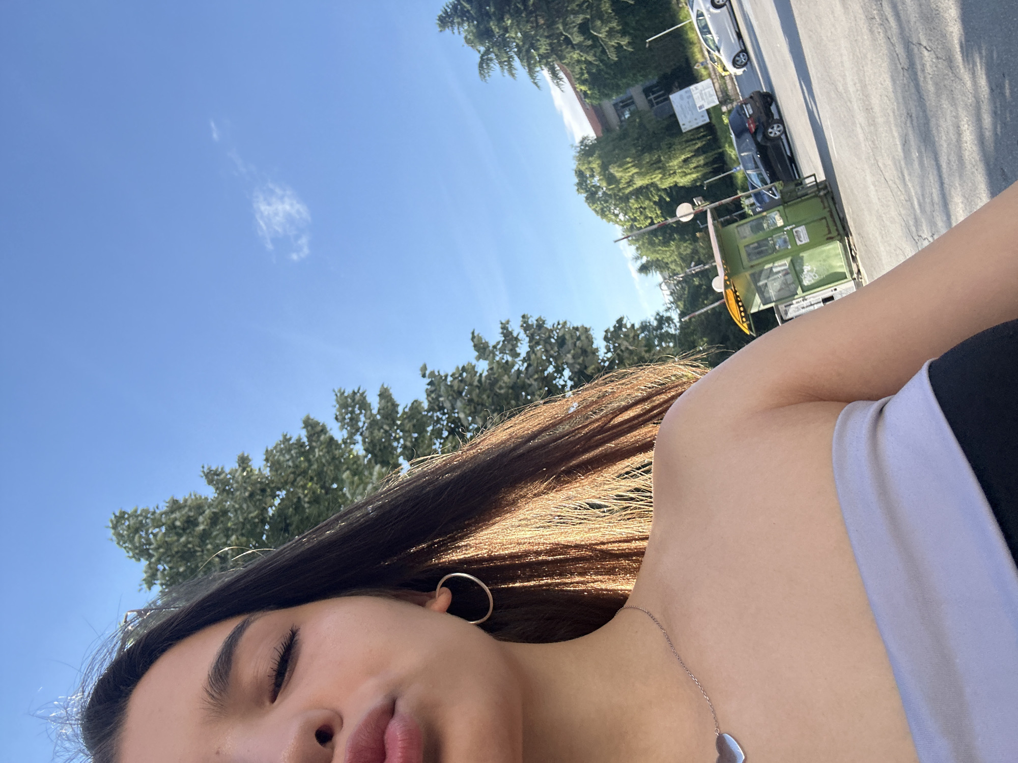 The image is a close-up, selfie-style photograph of a young woman taken outdoors in Shumen, Bulgaria, likely near a street. The woman is positioned on the left side of the frame, with her face partially visible. She has dark hair, and her eyes are closed, with her lips pursed as if blowing a kiss. She is wearing a silver hoop earring and a silver necklace with a heart-shaped pendant. Her attire appears to be a strapless top, with one shoulder exposed.
The background shows a bright blue sky with a wispy cloud. To the right of the woman, trees with green foliage are visible, some with dappled sunlight shining through. In the middle ground, there is a small green booth, possibly a security or ticket booth, with graffiti on its exterior. Several cars are parked or driving on an asphalt road in front of the booth. A building with a red roof is visible behind the trees in the distance. The lighting suggests it is daytime, with the sun casting distinct shadows. The overall impression is a casual, candid snapshot taken in an urban or semi-urban environment.