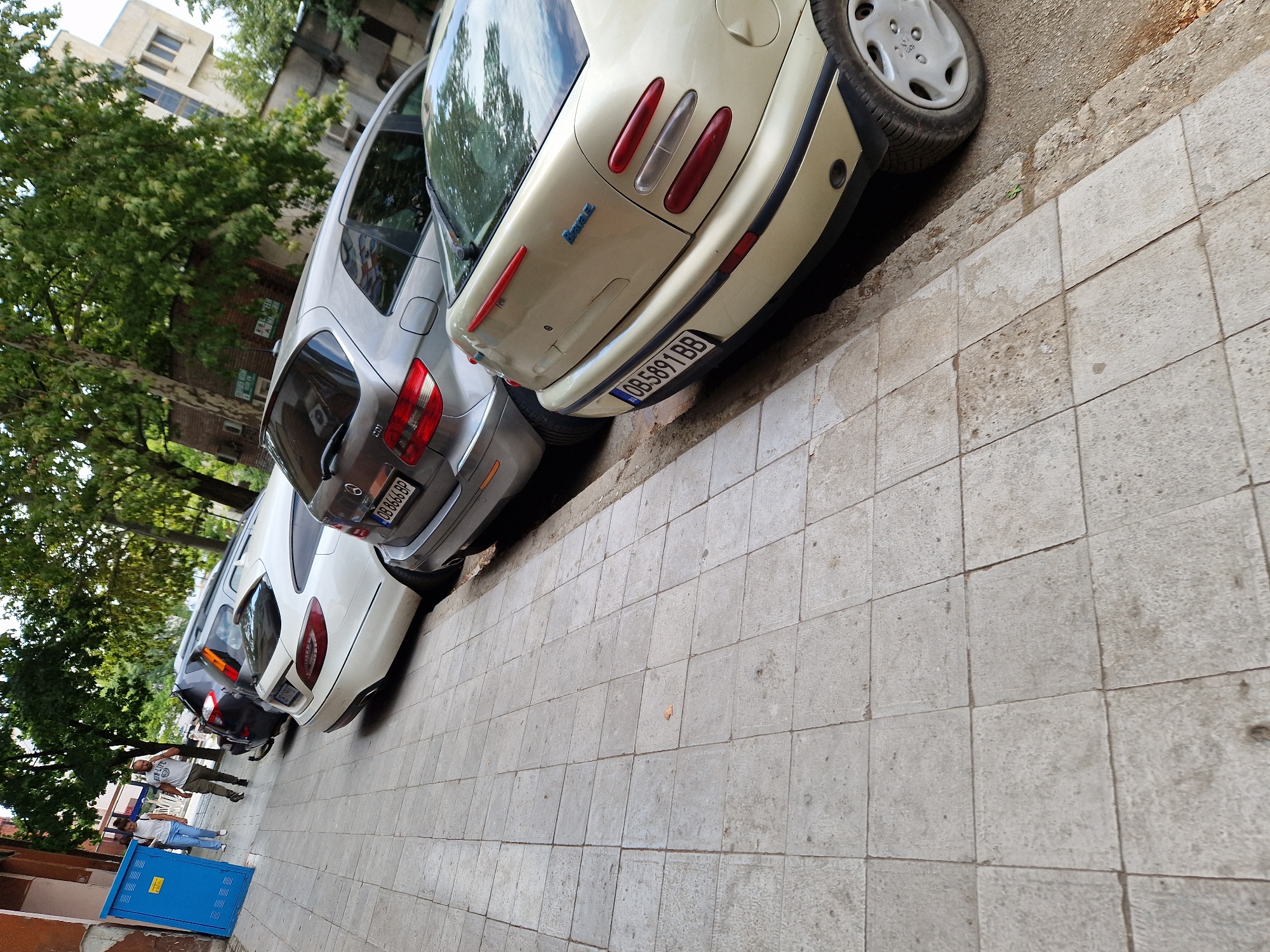 The image depicts a street scene in Lovech, Bulgaria, with several parked cars on the right side of a tiled sidewalk. The foreground is dominated by the tiled sidewalk and the rear of a light-colored Fiat Bravo. Beyond the Fiat, a silver Mercedes-Benz hatchback is parked, followed by a dark-colored SUV, and another Mercedes-Benz sedan. Two people are walking on the sidewalk in the background, near a blue utility box. The environment appears to be urban, with trees lining the street and buildings visible in the distance. The overall impression is of a typical day in a residential or commercial area of the city.