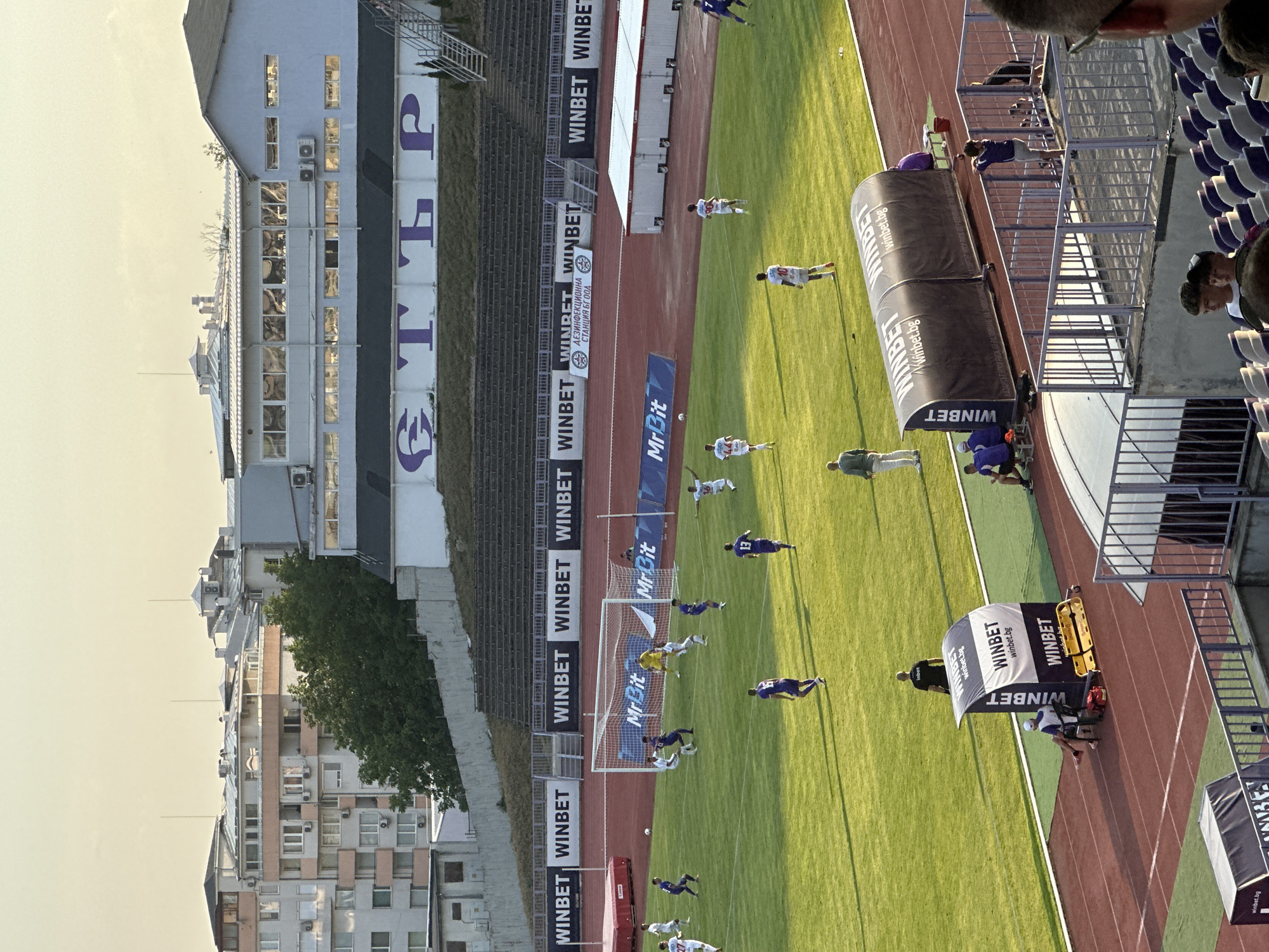 The image depicts a football match in progress at a stadium in Veliko Tarnovo, Bulgaria. The stadium is surrounded by buildings and trees. The field is green with white markings, and the goal is visible. Several players are on the field, some in action, others observing. Spectators are seated in the stands, and some are standing. Advertising banners are displayed around the stadium, including "WINBET." The sky is a pale yellow, suggesting it is either early morning or late afternoon. The overall atmosphere is one of a sporting event taking place in an urban environment.