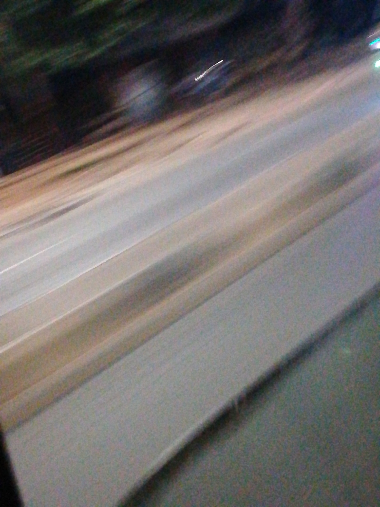 The image depicts a blurry, motion-blurred view of a road, possibly taken from a moving vehicle. The road surface is light-colored, with lane markings visible. The side of the road is lined with a sidewalk or shoulder, which is darker in color. On the left side of the image, there is some indistinct, dark vegetation or trees. There is no evidence of people or other subjects. The location in Lagos, Nigeria, cannot be determined from the image.