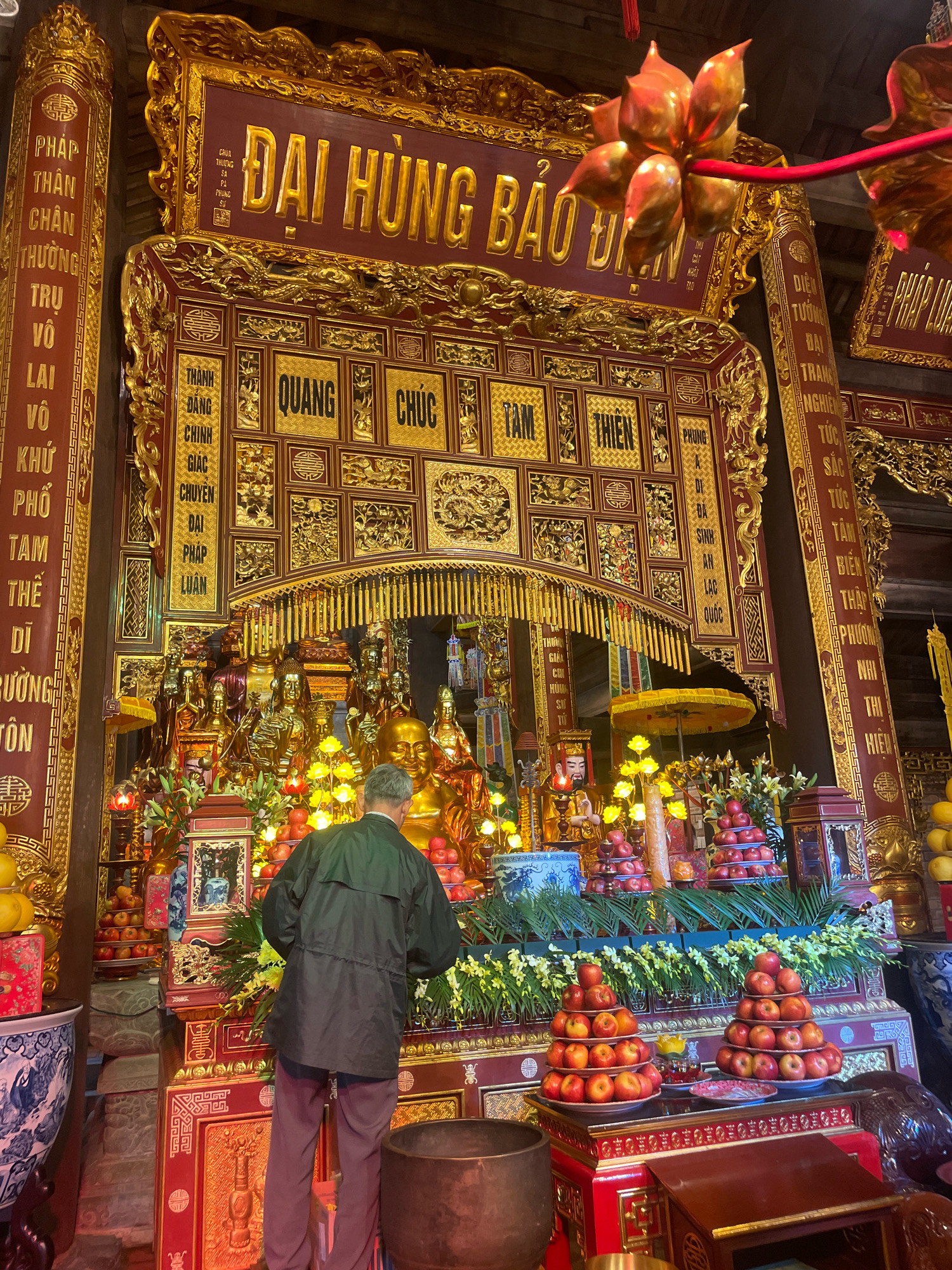 The image depicts the interior of a temple in Vietnam. A man in a dark green jacket and maroon pants is standing with his back to the camera, facing a large, ornate altar. The altar is adorned with numerous golden Buddhist statues, elaborate carvings, and decorative panels with text. Piles of red apples are stacked on plates at the front of the altar, alongside floral arrangements and other offerings. The background features red and gold architectural elements and more golden decorative details. The overall scene suggests a religious ceremony or a moment of prayer.