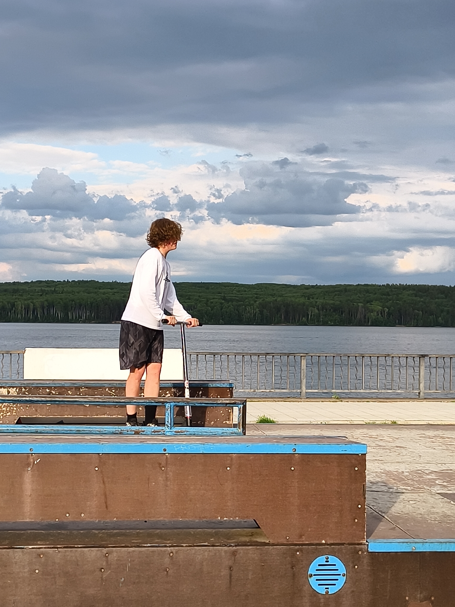 A lone male teenager with curly brown hair stands on a scooter on the edge of a skate park. He is wearing a long-sleeved white shirt and dark patterned shorts. The skate park features ramps made of wood with blue trim. In the background, a wide river stretches towards a dense forest of green trees. The sky above is filled with dramatic, dark clouds, suggesting the possibility of rain. A metal fence runs along the edge of the skate park, separating it from a paved area. The scene appears to be located in Chaikovsky, Russia, likely near the Kama River reservoir, which is bordered by extensive forests.