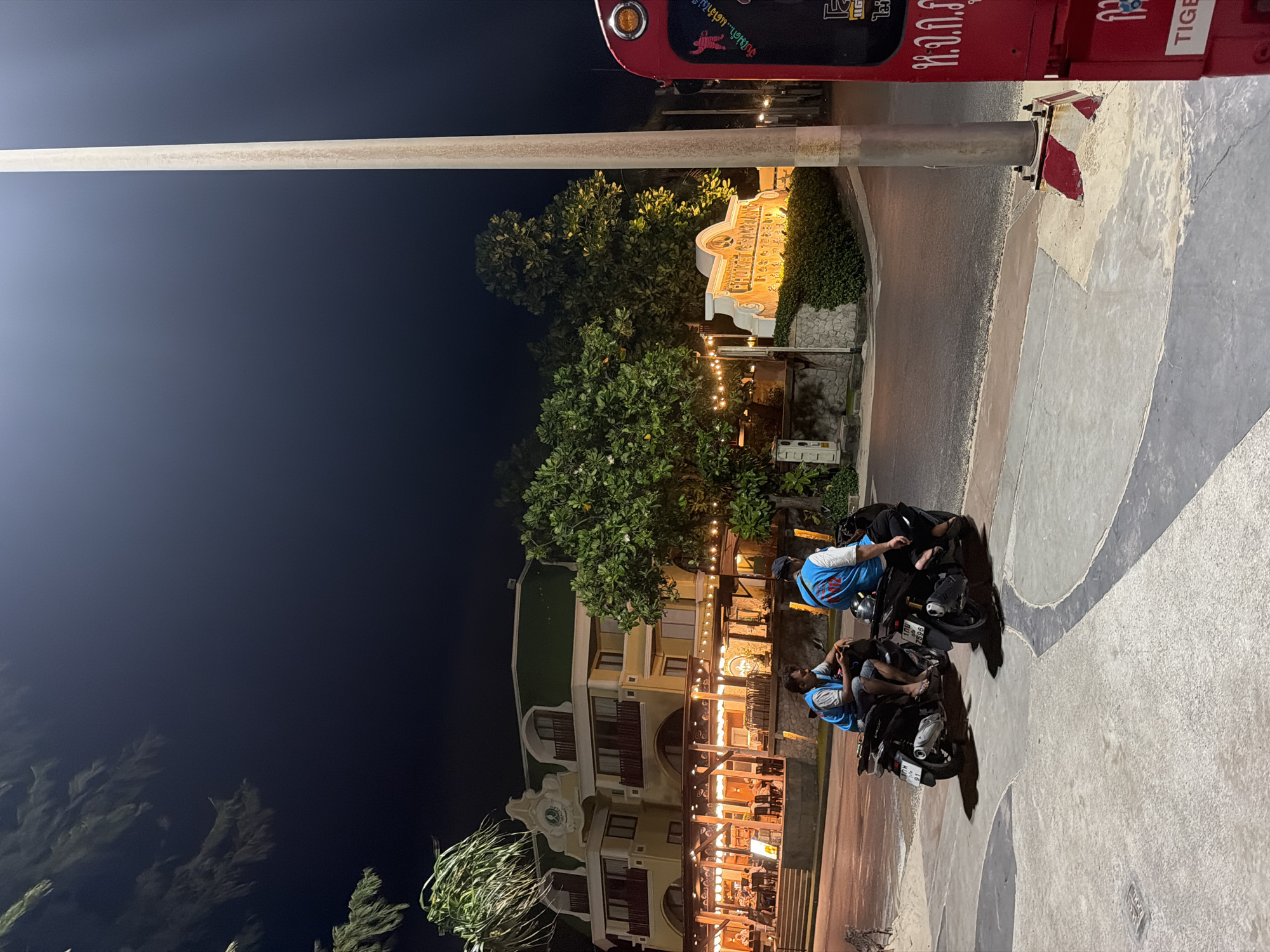 The image depicts a nighttime street scene in Patong, Thailand, likely near a beachside area. In the foreground, two men wearing blue vests are seated on motorbikes. The man on the left is looking at his phone, while the man on the right is facing forward. Both motorbikes are parked on a concrete pathway.
Behind them, a large, illuminated building with a distinctive yellow facade and a green roof is visible. The building has multiple floors and balconies, and its ground level appears to be a restaurant or hotel entrance with outdoor seating illuminated by string lights.
To the right of the building, a prominent sign with white lettering and an illuminated border reads "CHALONG COUNTRY CLUB." This sign is situated amidst lush green trees and shrubs. A tall, grey utility pole stands near the sign.
Further to the right, a portion of a red vehicle, possibly a tuk-tuk or a service vehicle, is visible, displaying Thai script. The overall environment suggests a lively urban or tourist area at night. The road surface is asphalt, and there is sand visible on the left side of the pathway.